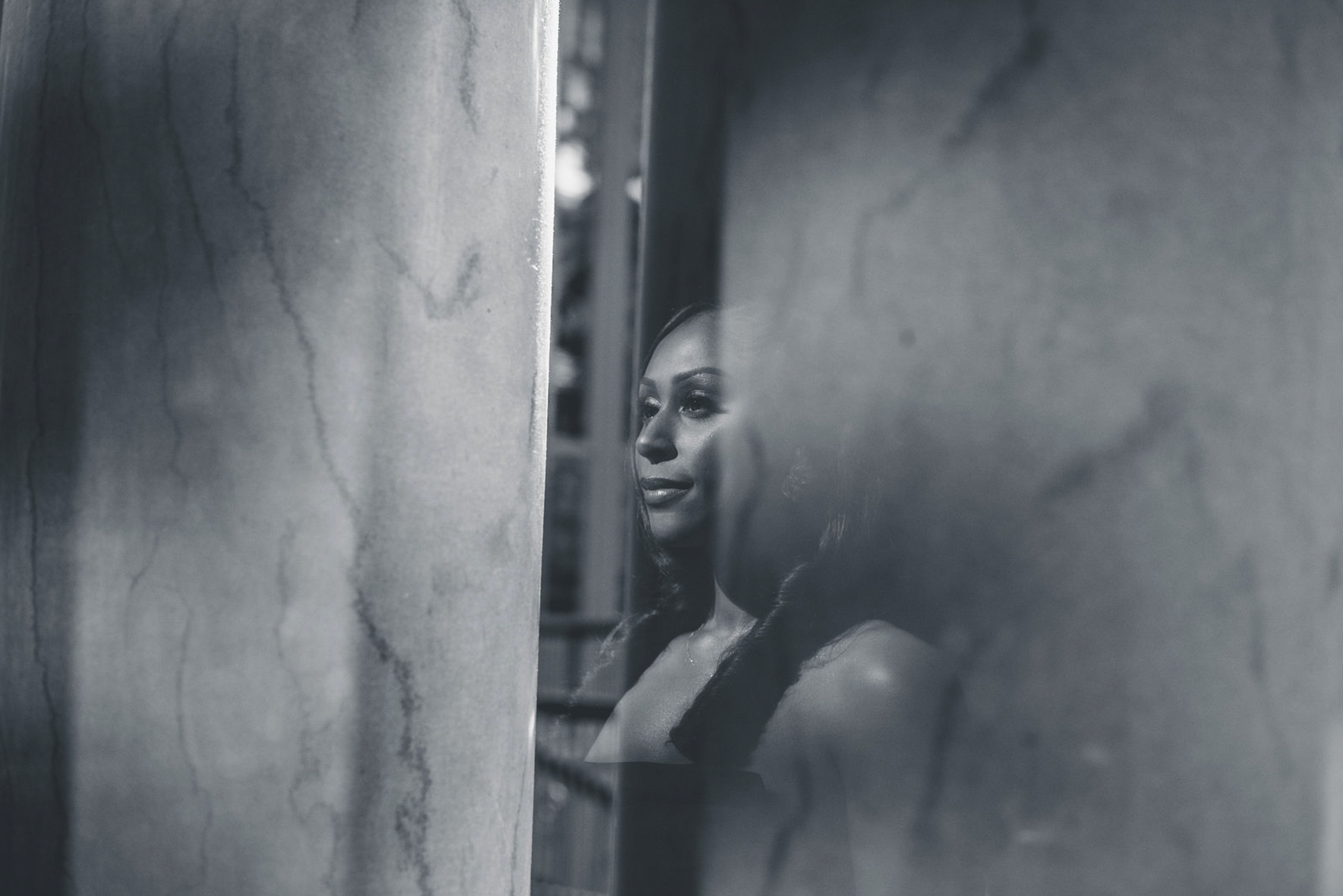 Black and white photograph of a woman with wavy hair looking through a narrow opening between two concrete walls.