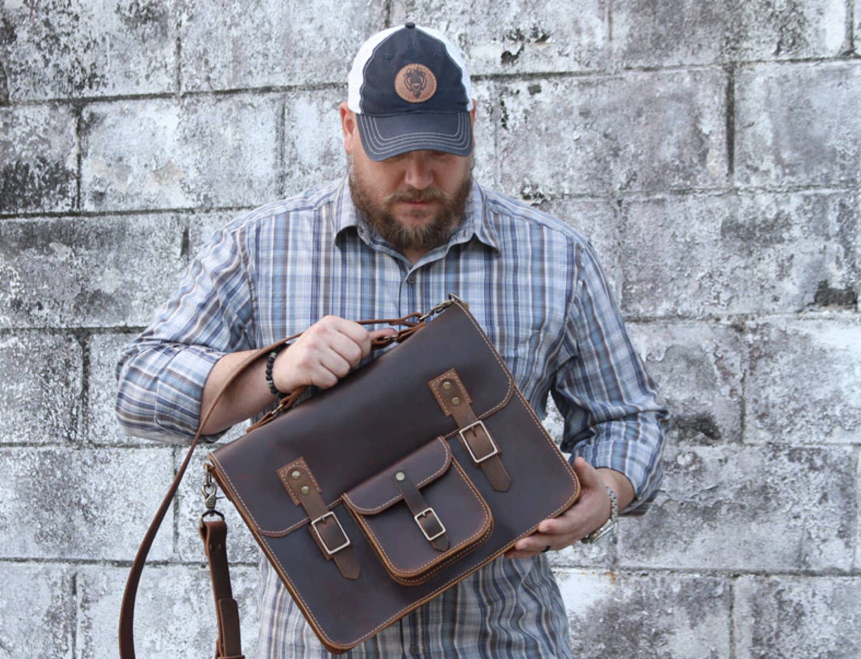 Man in plaid shirt with leather bag and cap standing against a concrete wall.