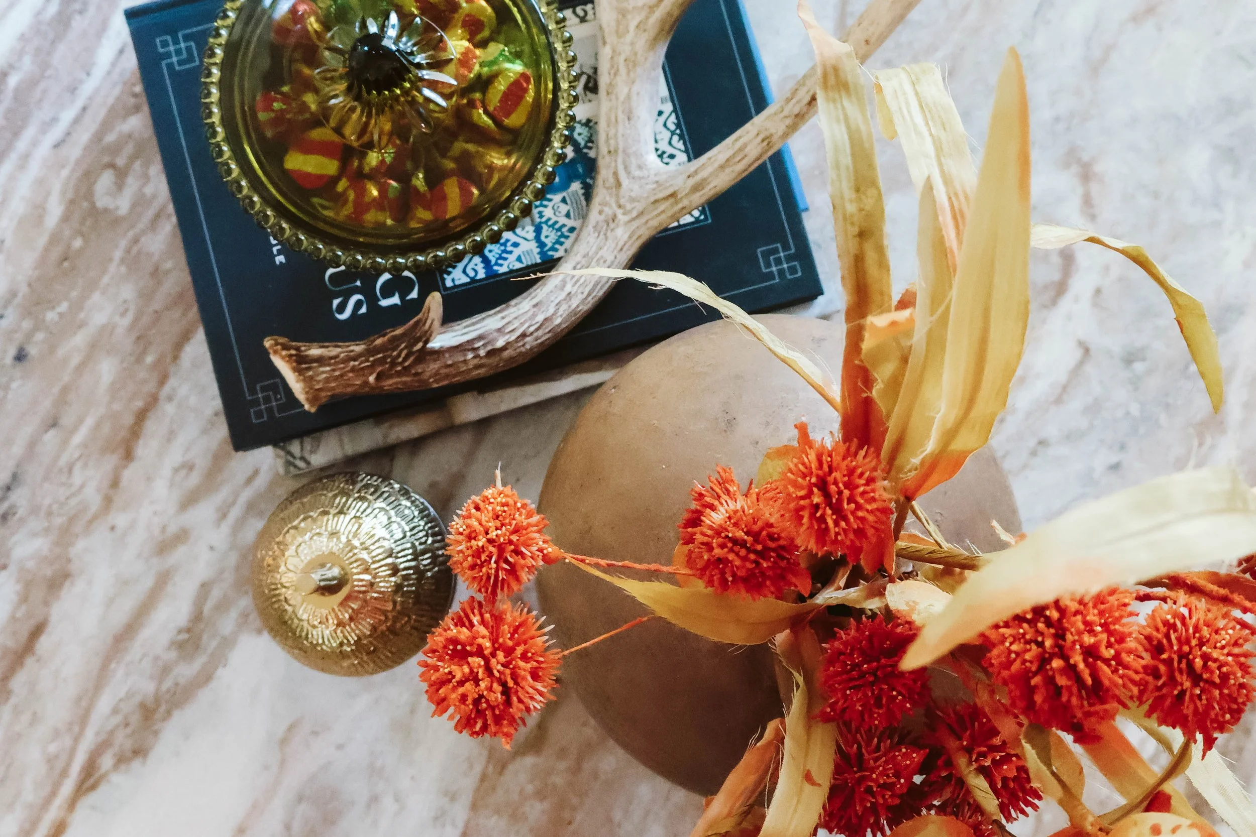 Decorative arrangement on a marble surface, featuring a bouquet of dried orange and yellow flowers, a small golden ornament, a beige round object, and a dark container with a gold lid containing multicolored wrapped candies, placed on top of a black book or magazine.