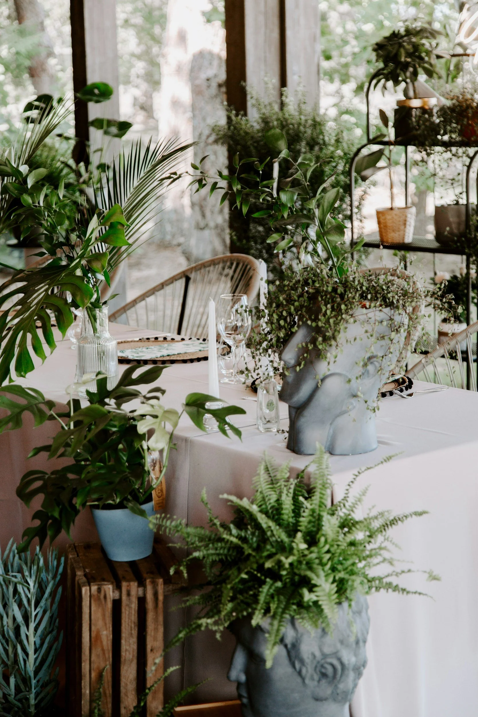 A dining table decorated with various green potted plants and a large sculpted head-shaped flower vase filled with greenery, set against a background of trees and a screened porch.
