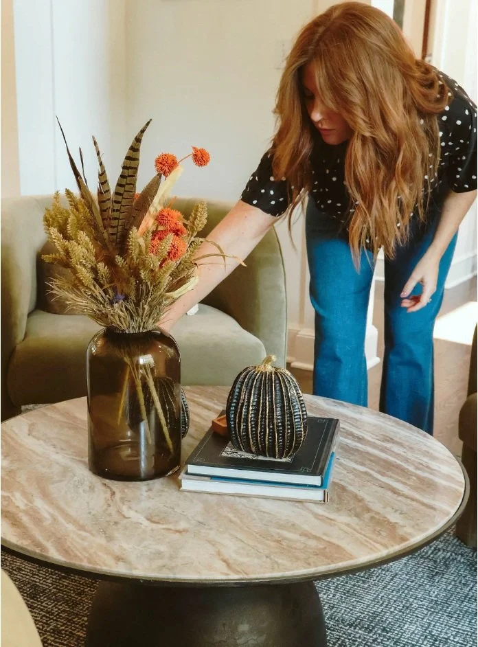 A woman with long red hair arranging a bouquet of dried flowers in a brown vase on a round marble coffee table, which also has a decorative pumpkin and a stack of books.