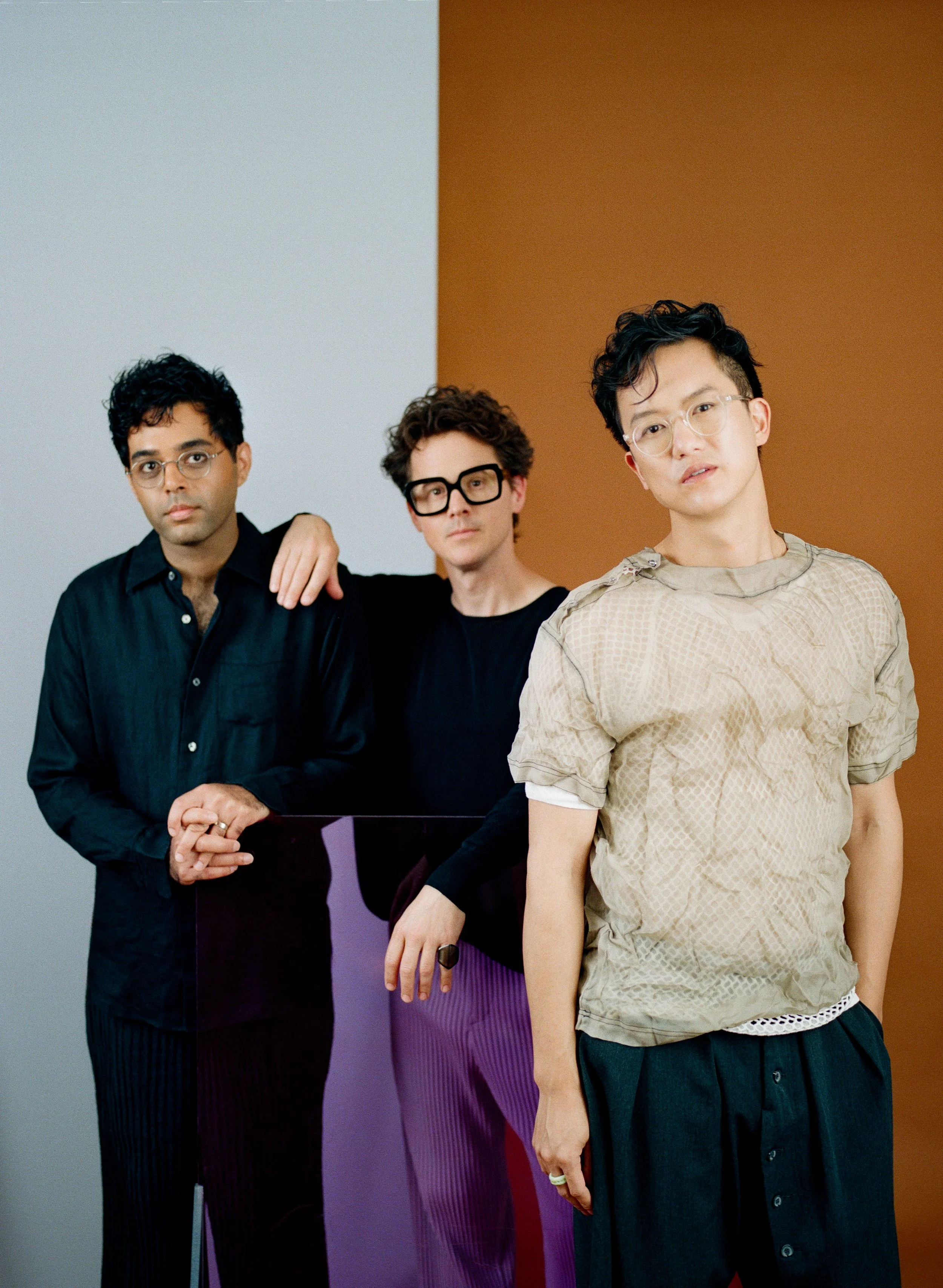 The band Son Lux standing against a background divided into sections of white, purple, red, and brown, with one person on the right in the foreground and the other two partially behind. They are wearing casual clothing and glasses.