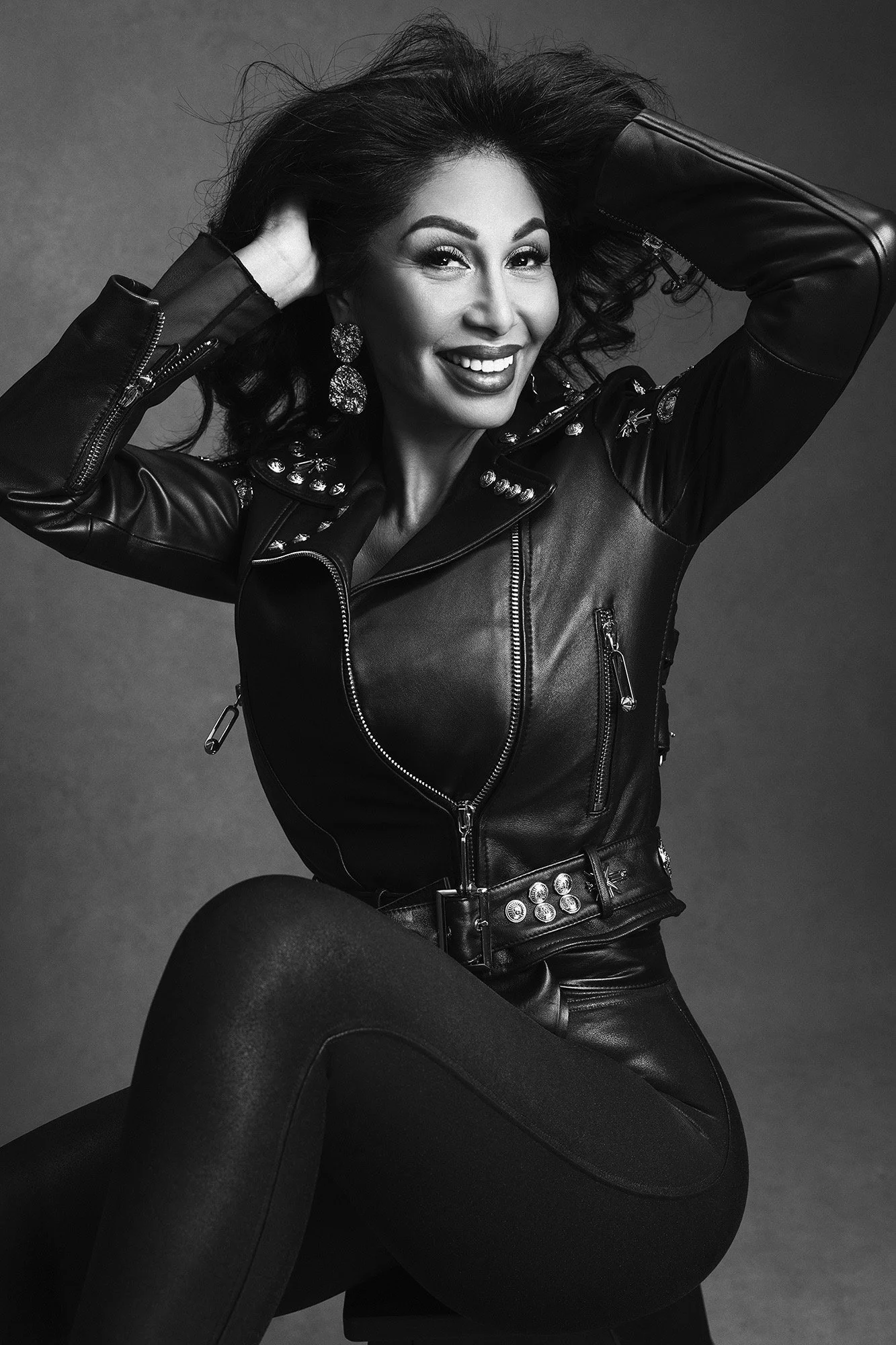 Eine Frau über 40 in einer Lederjacke mit Nieten, die Hände im wilden dunklen Haar, lacht mit ungezähmter Freude und Energie — Editorial-Portrait — 40 Über 40 Projekt, fotografiert von Mayumi Acosta Photography, Fotostudio Innsbruck, Tirol.