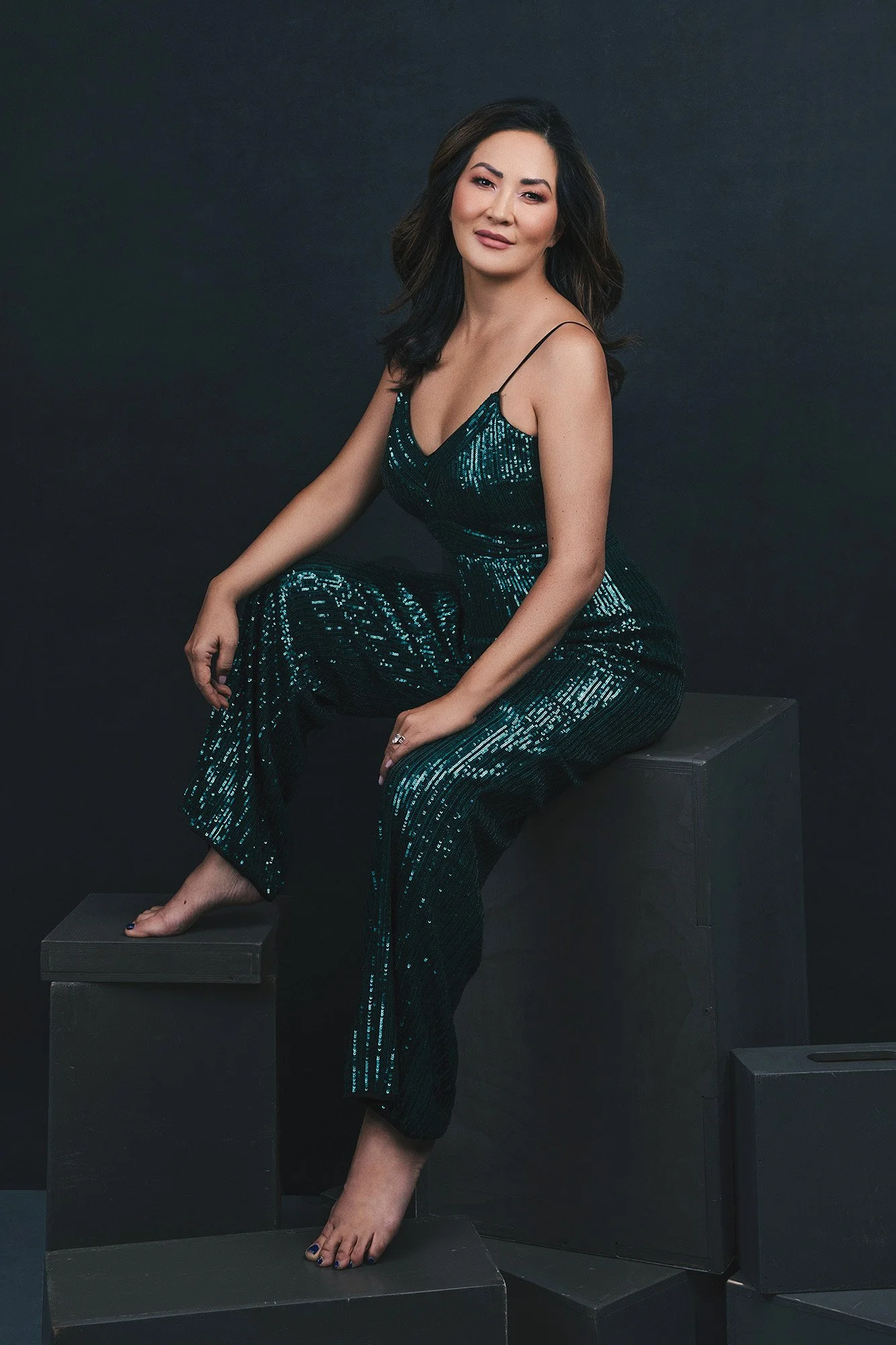 Vogue Style Portrait einer Frau über 40 im türkisfarbenen Paillettenoverall, barfuß auf Studio-Würfeln sitzend mit müheloser Eleganz und leuchtendem Blick — iconic woman portrait series von Mayumi Acosta Photography, Fotostudio Innsbruck, Tirol.