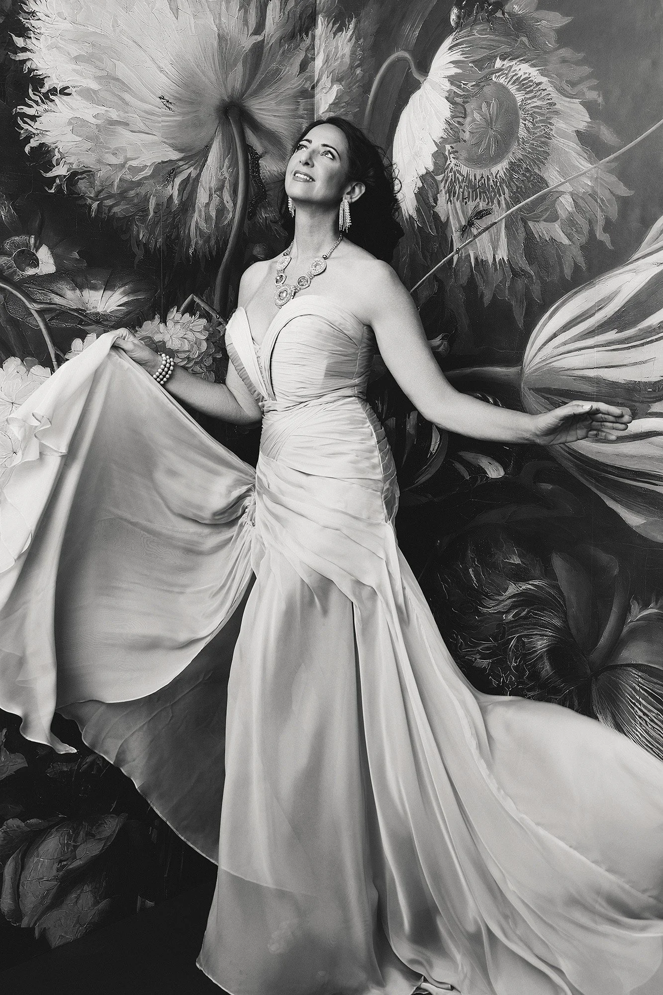A woman over 40 in a flowing strapless gown lifting her skirt, gazing upward with wonder against a dramatic painted floral backdrop — fine art portrait — photographed by Mayumi Acosta Photography, photo studio Innsbruck, Tirol.