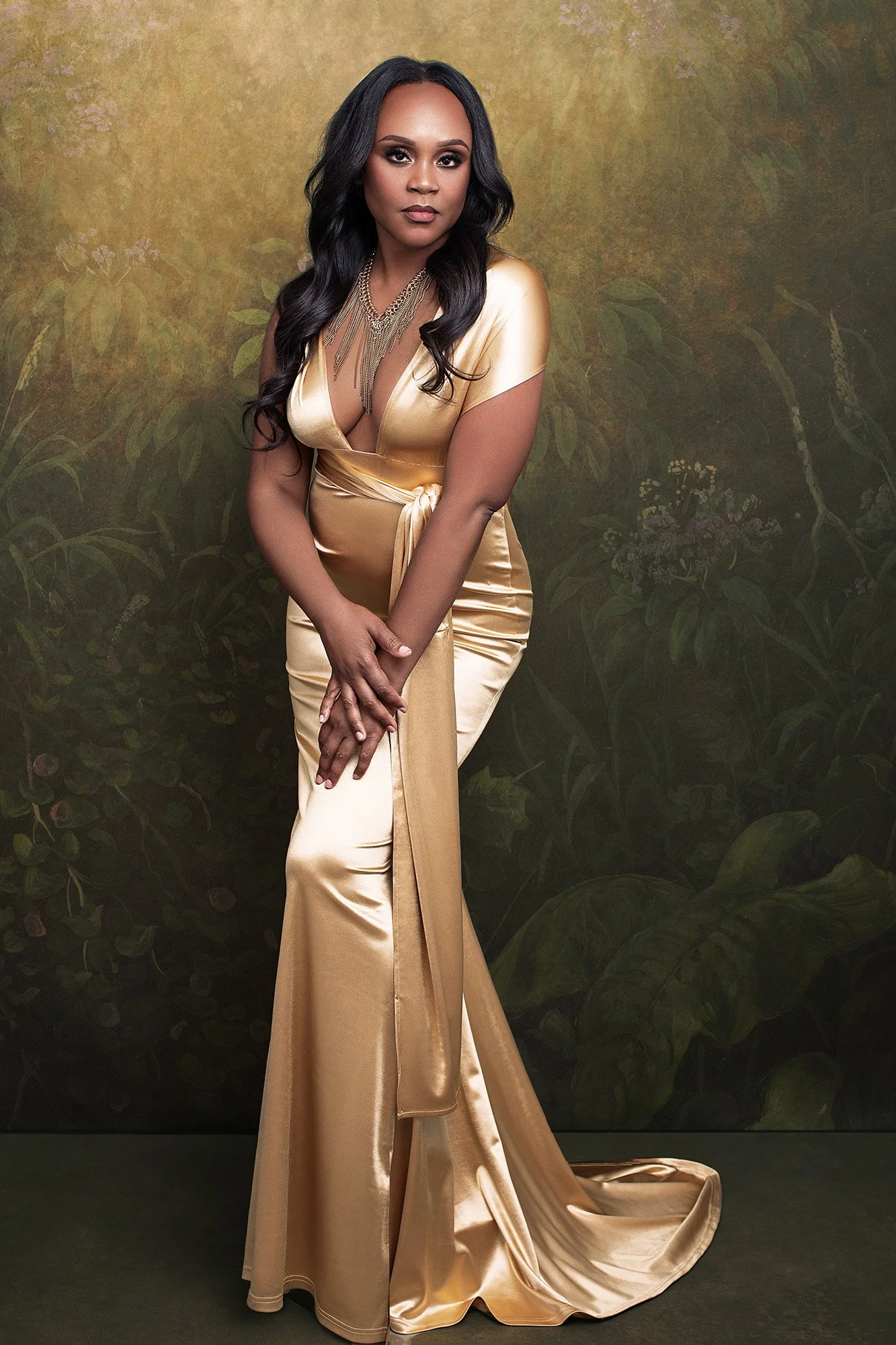 Luxus Fotoshooting Frau über 40 im goldenen Satinkleid mit Kettenhalskette, steht mit gefalteten Händen und souveräner Haltung vor botanischer Kulisse — 40 Über 40 Fotoprojekt von Mayumi Acosta Photography, Fotostudio Innsbruck, Tirol.