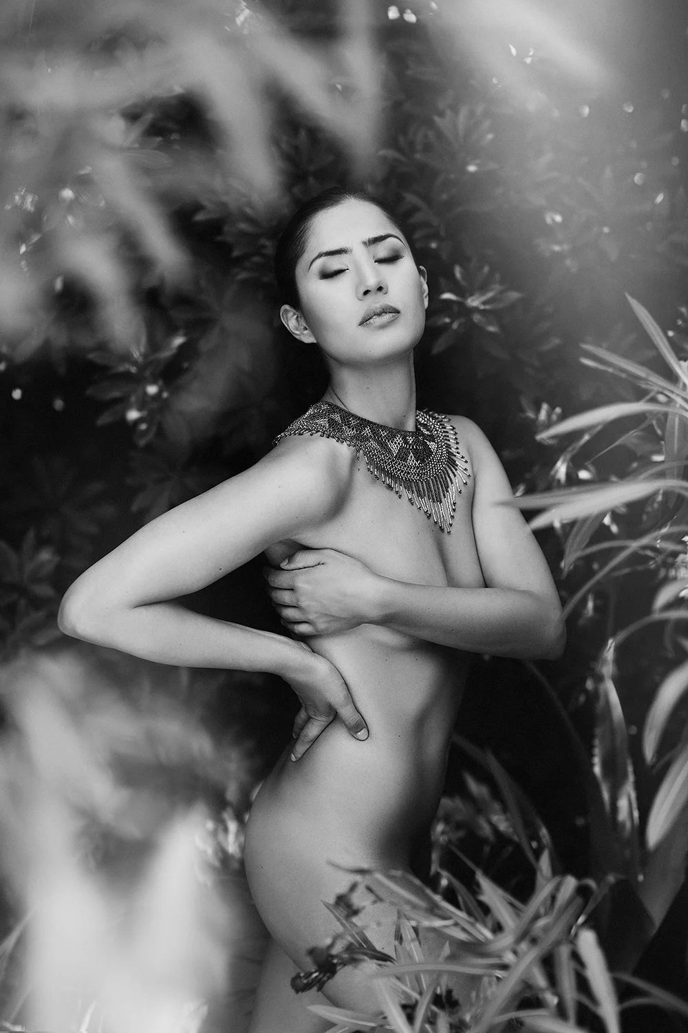 Fine art ethereal black and white nude portrait of a woman among botanical leaves with statement necklace — dreamlike artistic photography by Mayumi Acosta, Innsbruck Austria