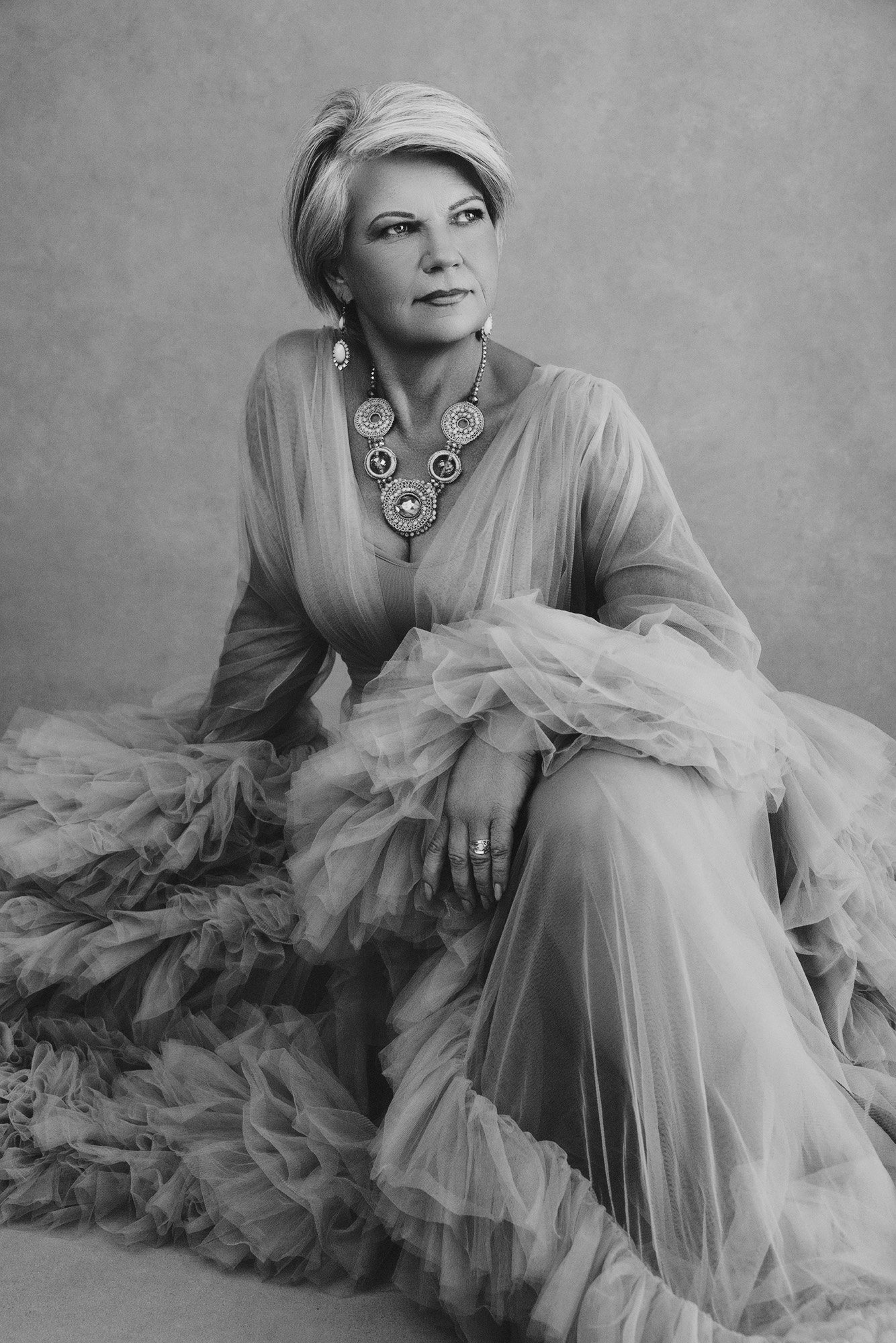 A woman over 40 seated in a flowing ruffled tulle gown, bold statement necklace, her gaze turned with quiet authority — elegant fine art portrait —iconic woman portrait series, photographed by Mayumi Acosta Photography, photo studio Innsbruck, Tirol.