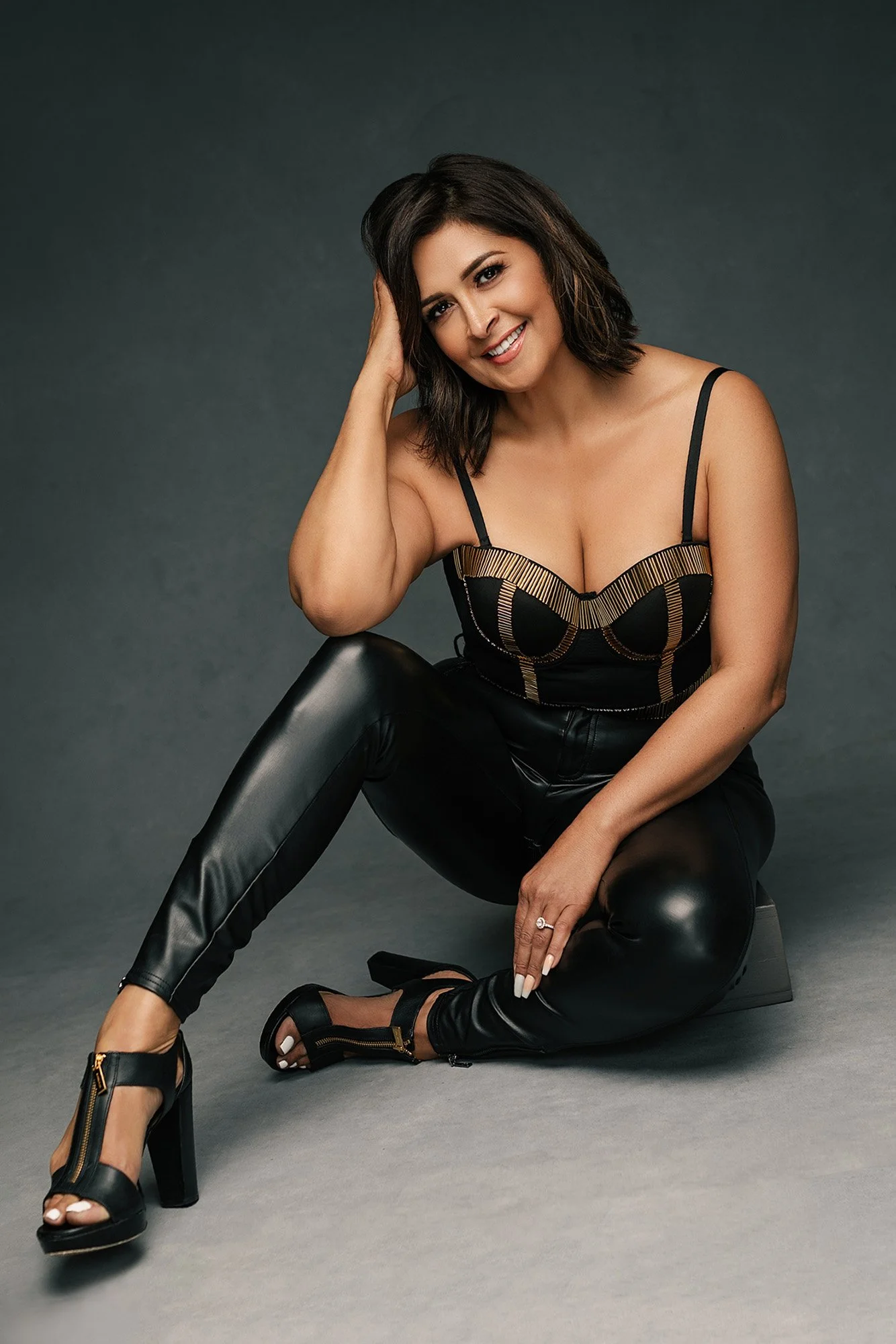 Moderne Glamour Fotografie Frauen über 40 — Frau in schwarzem und goldenem Korsett-Bustier mit Lederhose, auf dem Studioboden sitzend, Hand an der Schläfe, lächelt mit natürlicher Wärme und modernem Charme — iconic woman portrait series von Mayumi Ac