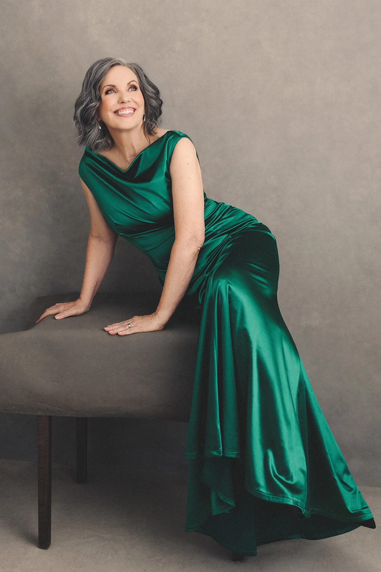 Luxus Fotoshooting Frau über 40 mit eleganten Silberwellen im smaragdgrünen Satinkleid, lehnt anmutig auf einer Samtbank und lacht voller Freude — 40 Über 40 Fotoprojekt von Mayumi Acosta Photography, Fotostudio Innsbruck, Tirol.