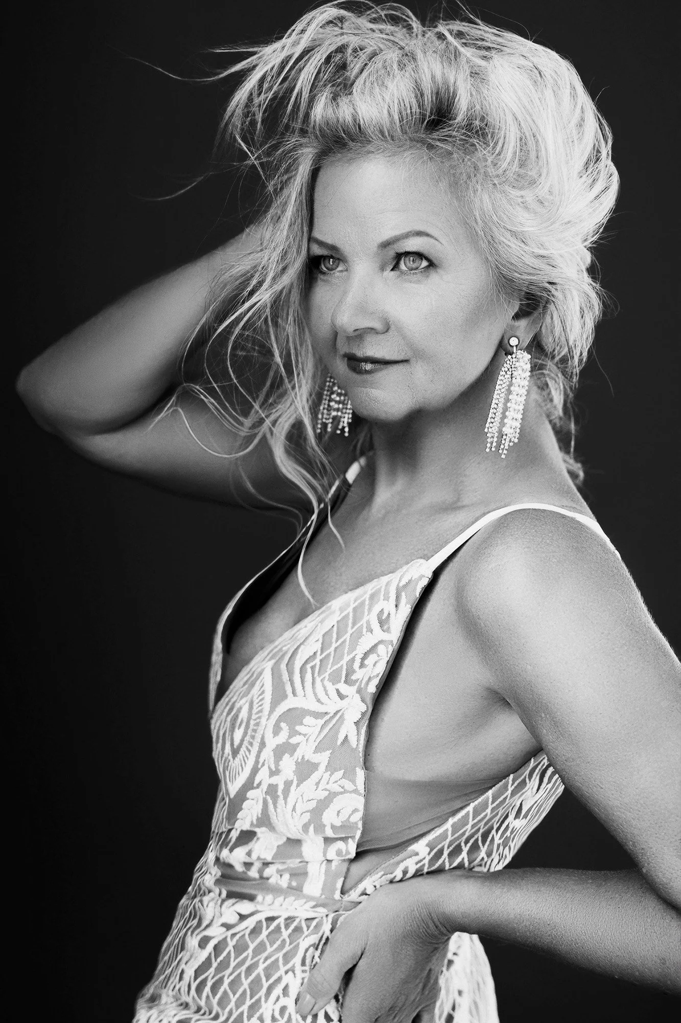 Glamour meets editorial portrait photography — a woman over 40 in an embroidered gown, hand in windswept blonde hair, looking over her shoulder with fierce magnetic energy — iconic woman portrait series by Mayumi Acosta Photography, photo studio Inns