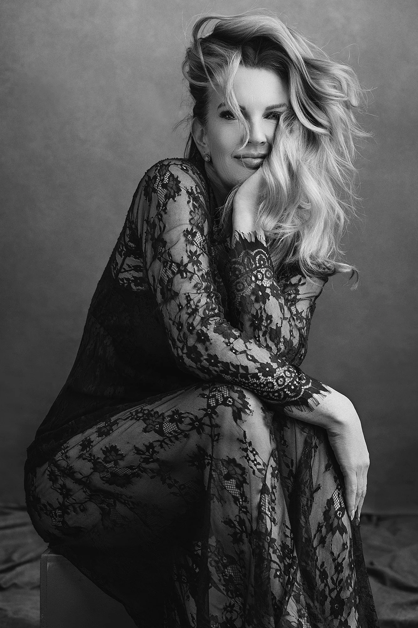 Hochwertiges Studio Portraitshooting Frau über 40 in transparentem schwarzen Spitzenkleid, mit verwehtem blonden Haar, Wange auf die Hand gestützt — zeitlose Sinnlichkeit und stille Mystik — iconic woman portrait series von Mayumi Acosta Photography,