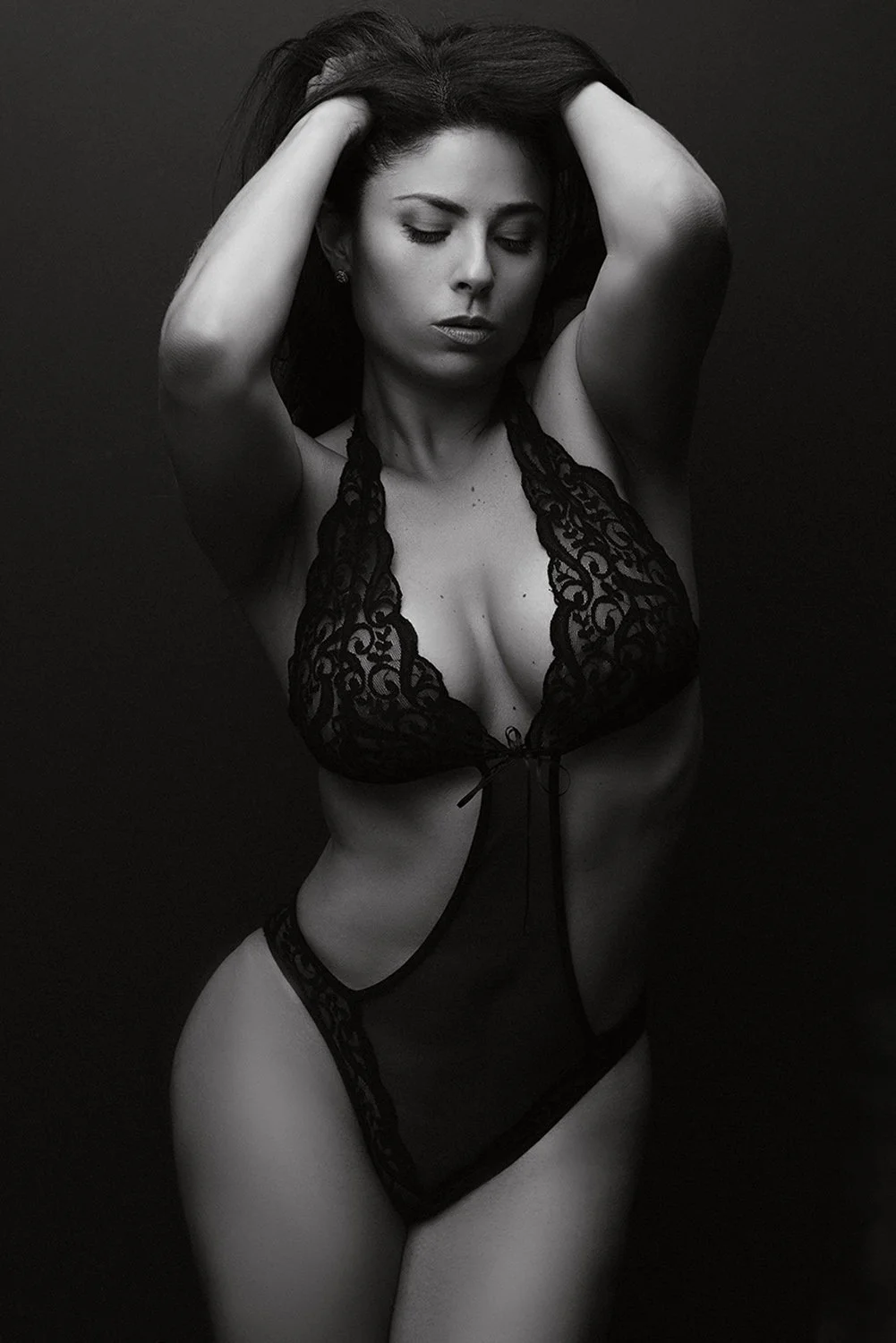 Fine art black and white boudoir portrait of a woman in black lace bodysuit with arms raised against a dark background — sculptural intimate photography by Mayumi Acosta, Innsbruck Austria