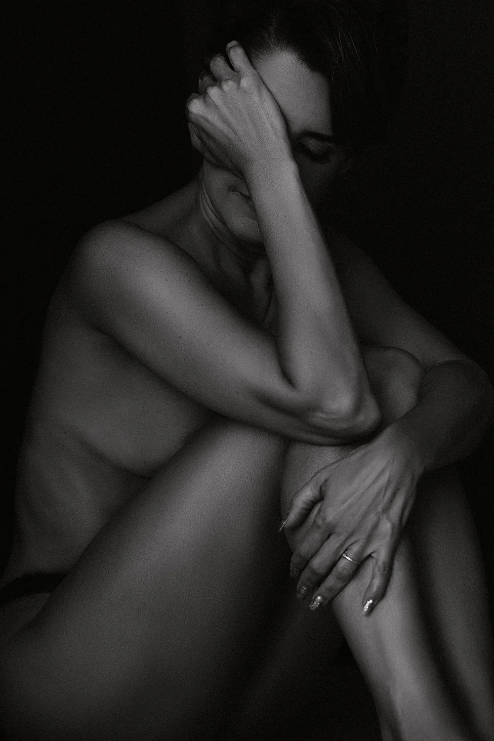 Fine art black and white intimate portrait of a woman in self-embrace against a dark background — deeply personal nude photography by Mayumi Acosta, Innsbruck Austria