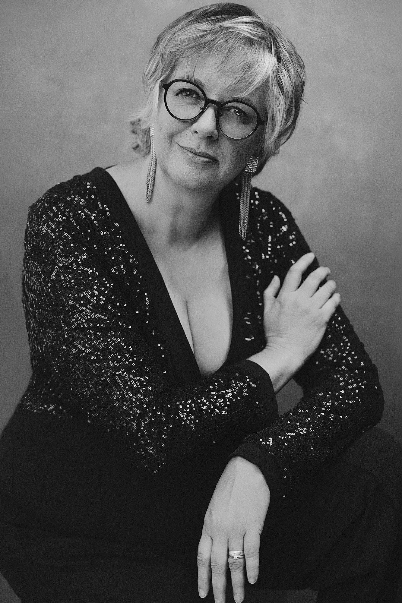 Style magazine portrait: A woman over 40 wearing a black sequined top with a plunging neckline, round glasses, and long crystal earrings; her gaze is calm and confident — “40 Over 40” photo project by Mayumi Acosta Photography, photo studio in Innsbr
