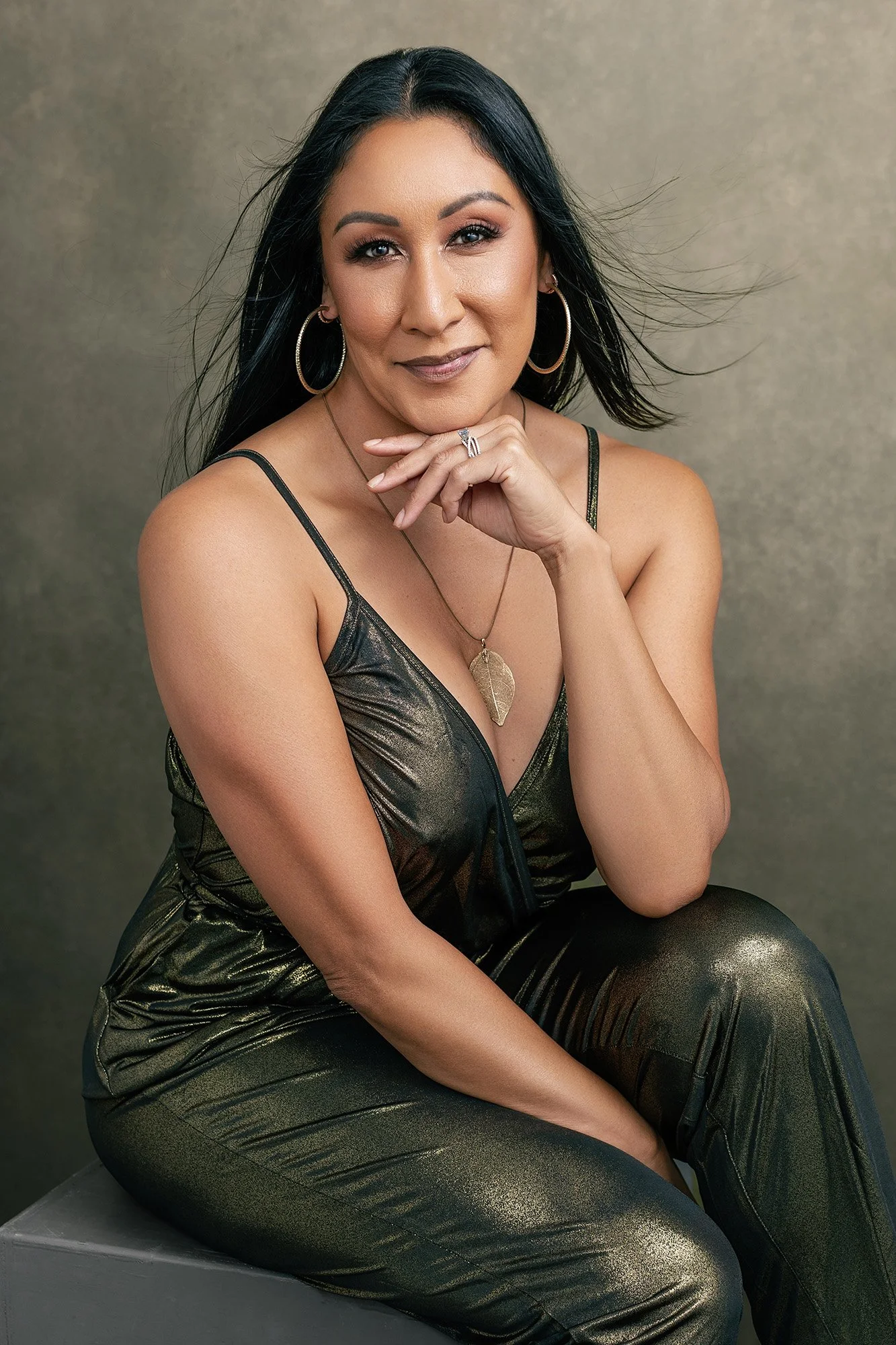 Modern glamour and fashion portrait session for women over 40 — a woman in a gold metallic jumpsuit, dark hair windswept, hand resting under her chin with a knowing smile and magnetic presence —portrait by Mayumi Acosta Photography, photo studio Inns