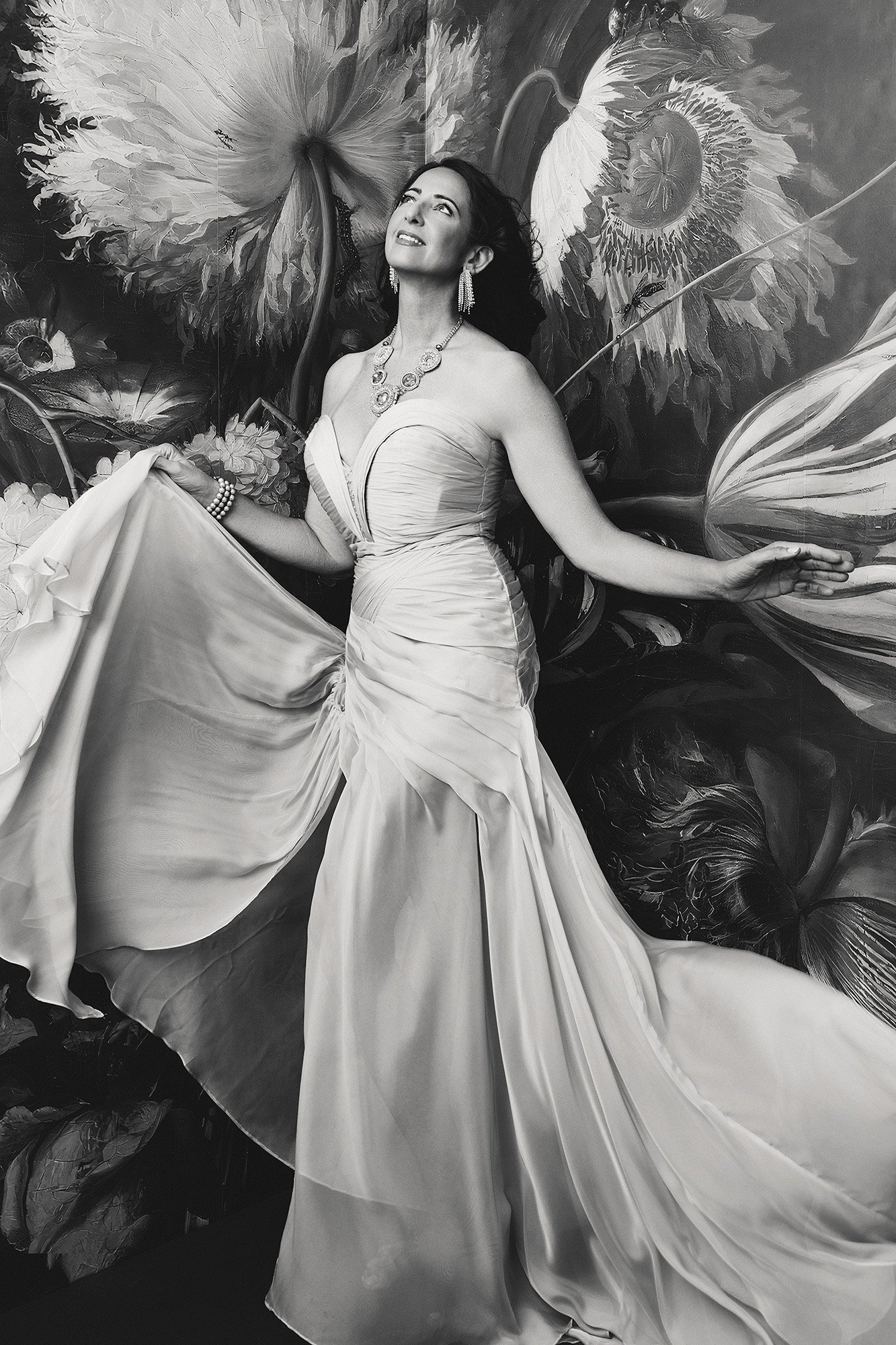 A woman over 40 in a flowing strapless gown lifting her skirt, gazing upward with wonder against a dramatic painted floral backdrop — fine art portrait — 40 Over 40 Project, photographed by Mayumi Acosta Photography, photo studio Innsbruck, Tirol.