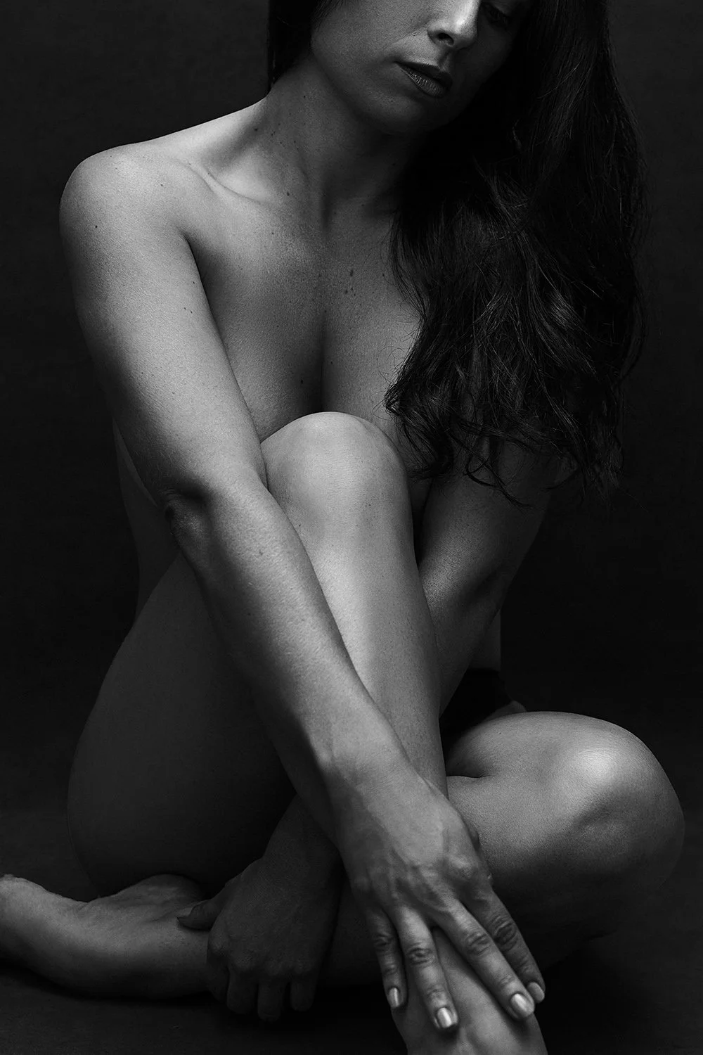 Fine art sculptural black and white nude portrait with interlocking arms and legs against a dark background — contemporary intimate photography by Mayumi Acosta, Innsbruck Austria