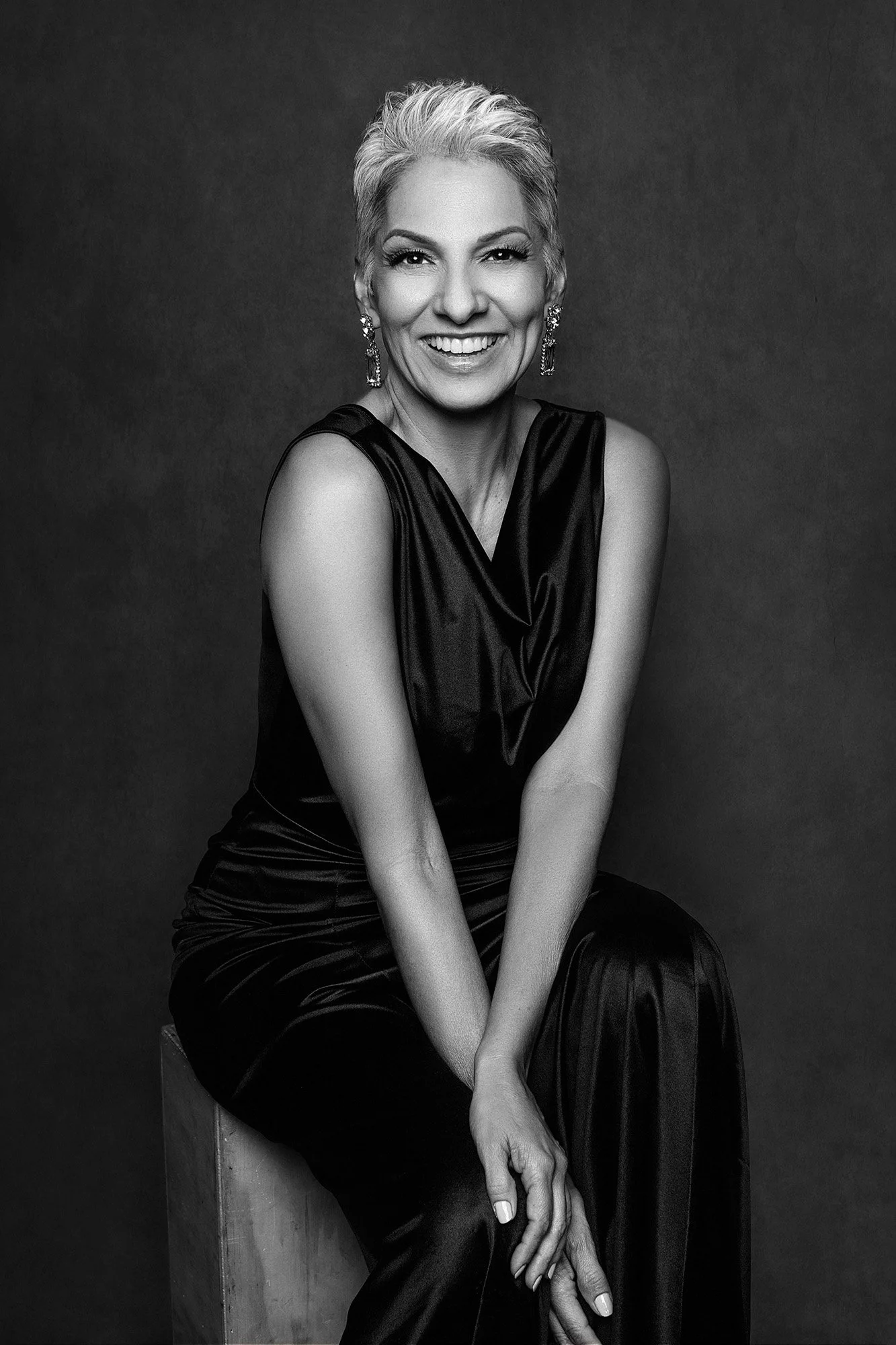 Magazine style fashion photography for women over 40 — a woman in a black sequin blazer with a statement fringe necklace, short blonde hair, seated with her gaze turned with cool, compelling authority —iconic woman portrait series by Mayumi Acosta Ph