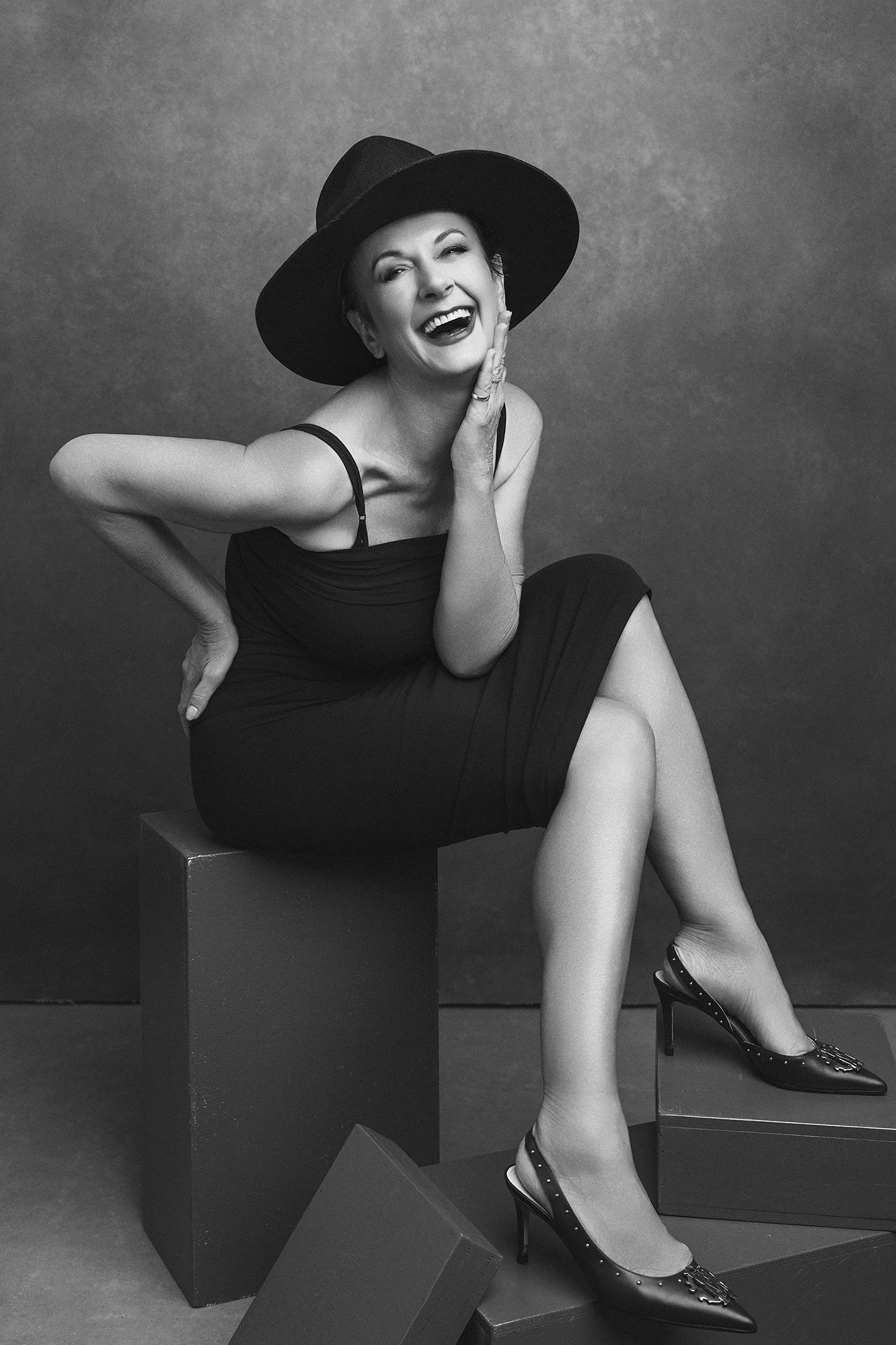 Magazin Style Portrait Frau über 40 im schwarzen Kleid und Hut, auf Studio-Würfeln sitzend, lacht mit ungezügelter Freude — iconic woman portrait series Mayumi Acosta Photography, Fotostudio Innsbruck, Tirol.