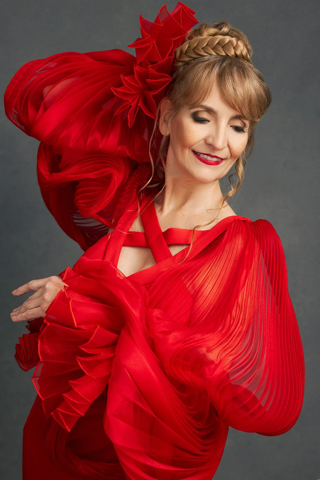 Vogue style portrait of a woman over 40 in a sculpted red pleated couture gown with floral headpiece, eyes downcast with a serene smile — fashion portrait photography for women Innsbruck Austria — iconic woman portrait series Mayumi Acosta Photograph