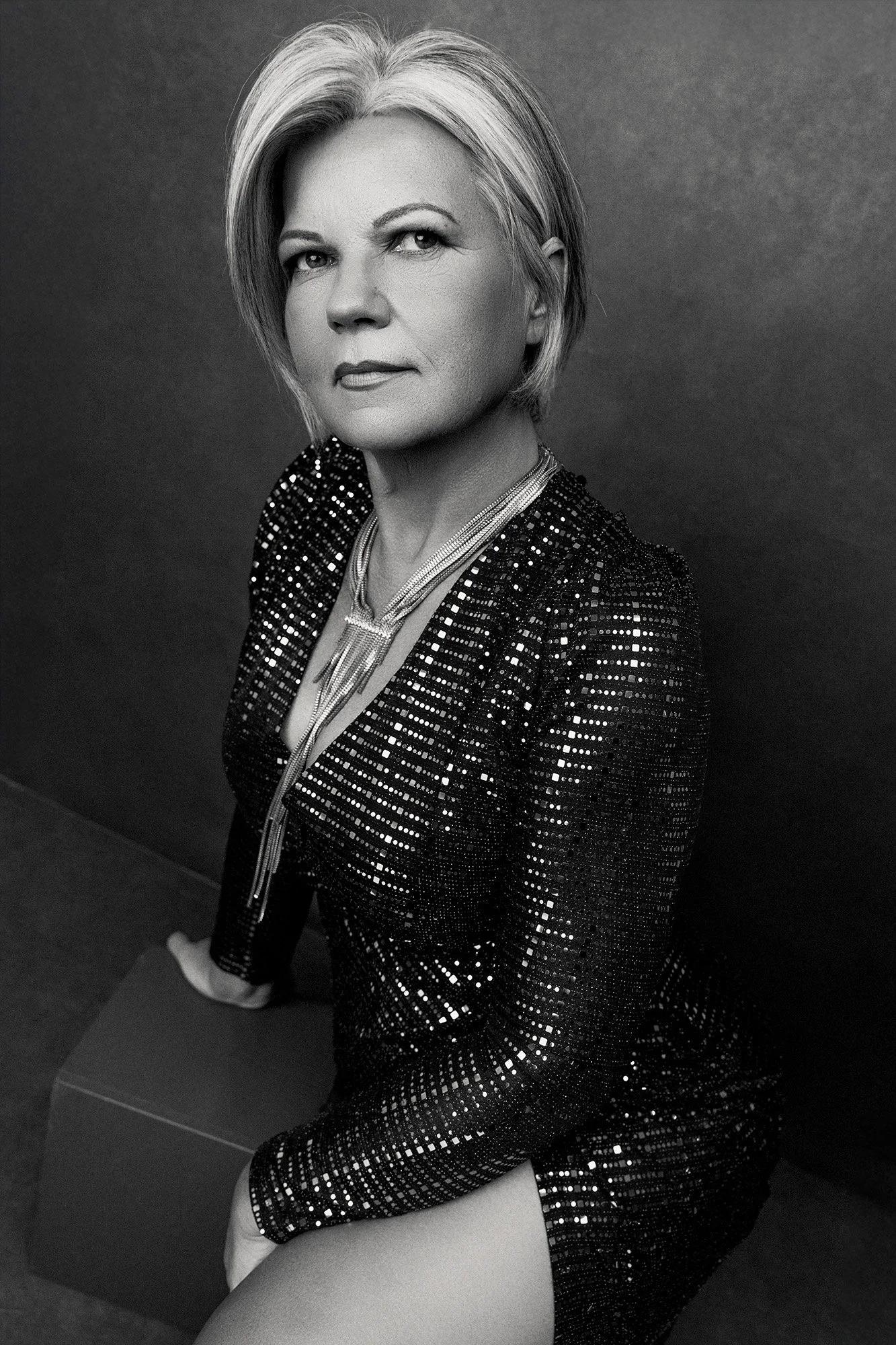 Magazine style fashion photography for women over 40 — a woman in a black sequin blazer with a statement fringe necklace, short blonde hair, seated with her gaze turned with cool, compelling authority —iconic woman portrait series by Mayumi Acosta Ph