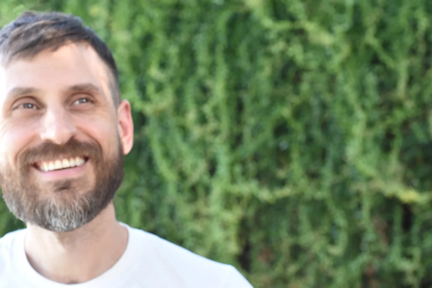 A happy man with a beard smiling outdoors against a green leafy background.