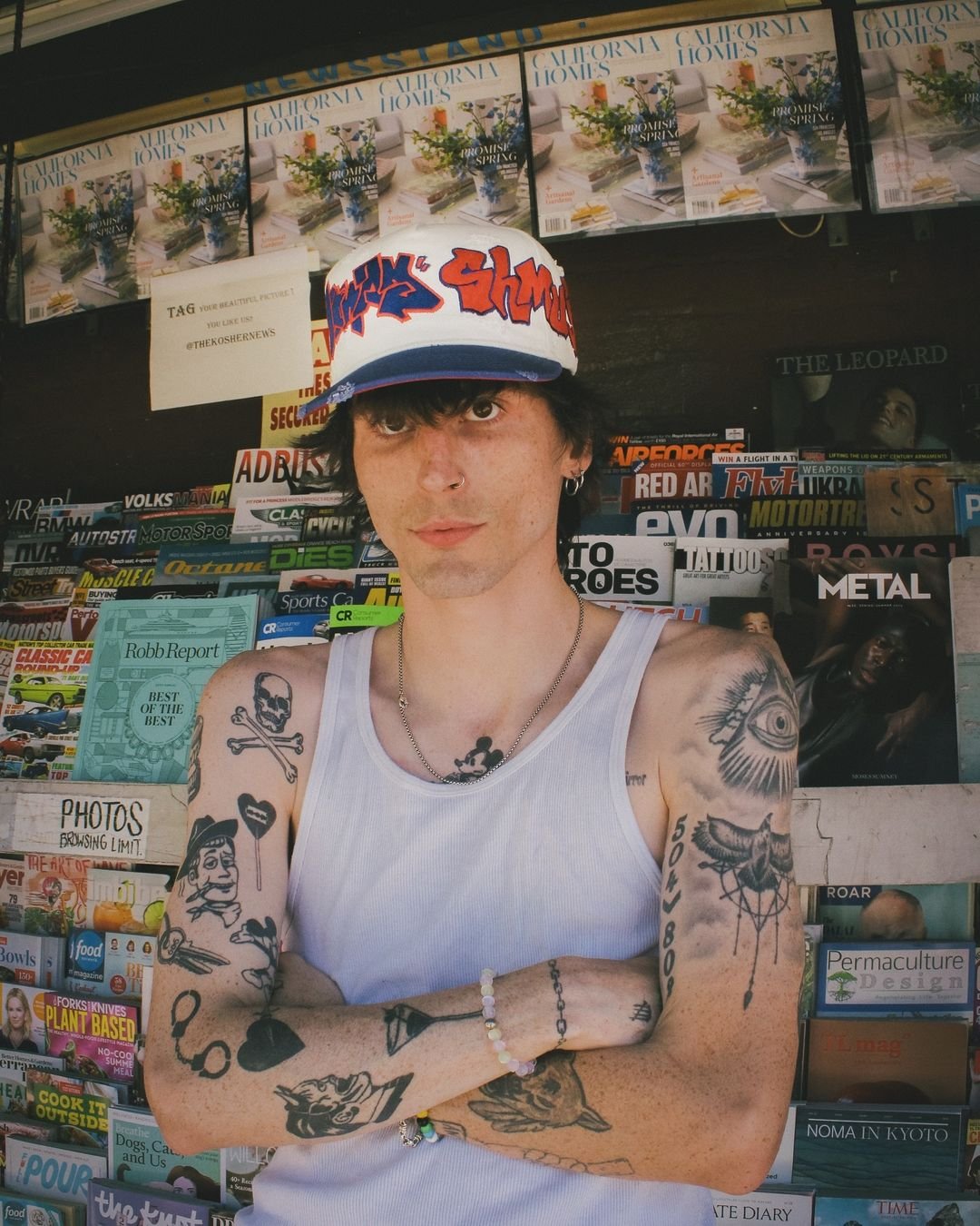 conswank standing in front of a magazine stand with his arms crossed, wearing a white beater tank top and a hat.