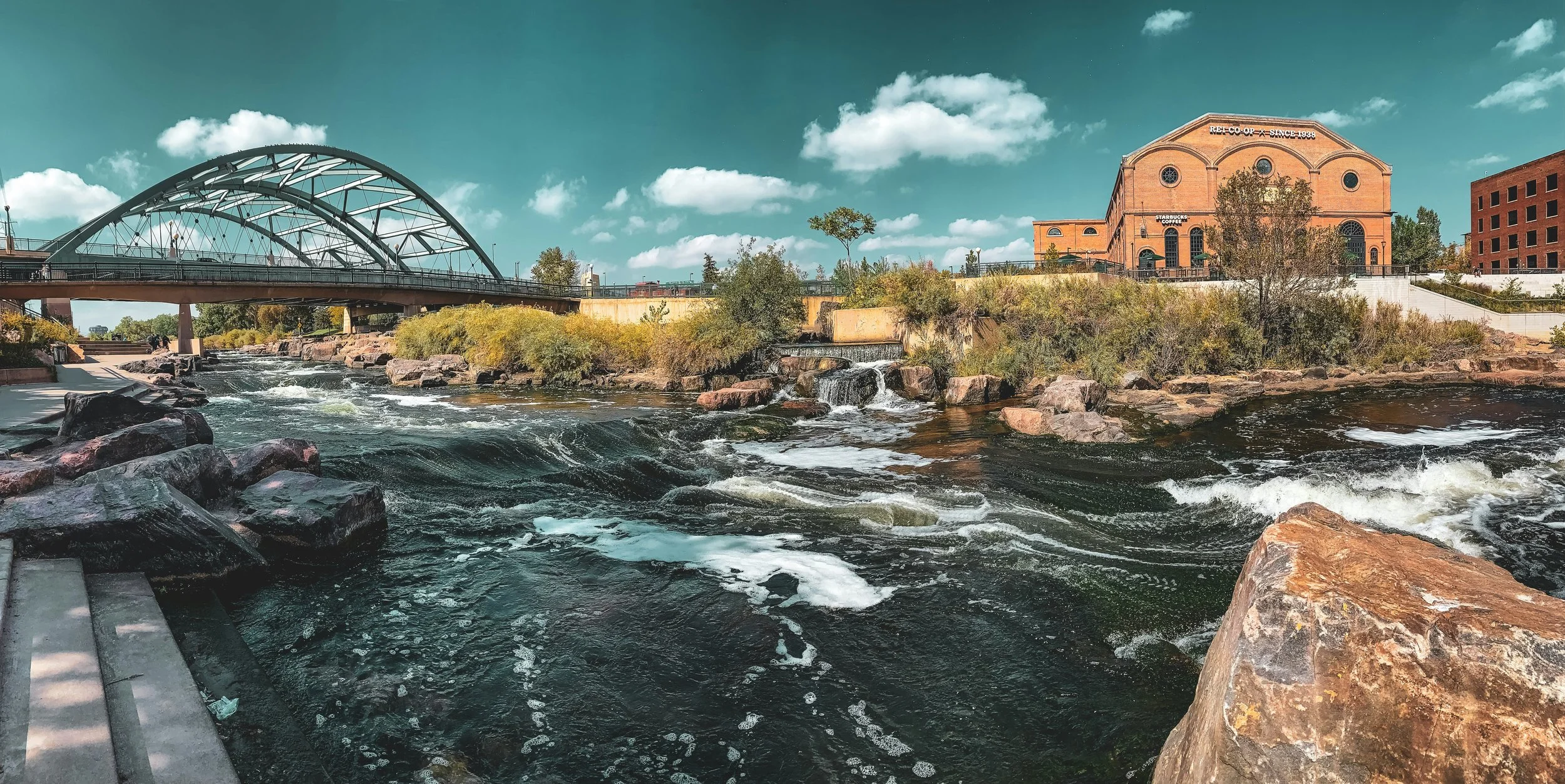 A scenic view of a river with rocks and small waterfalls, lined by a walkway and greenery. In the background, there is a bridge and a brick building with a sign reading 'REI CO-OP SINCE 1938' with a Starbucks Coffee sign, under a partly cloudy sky.