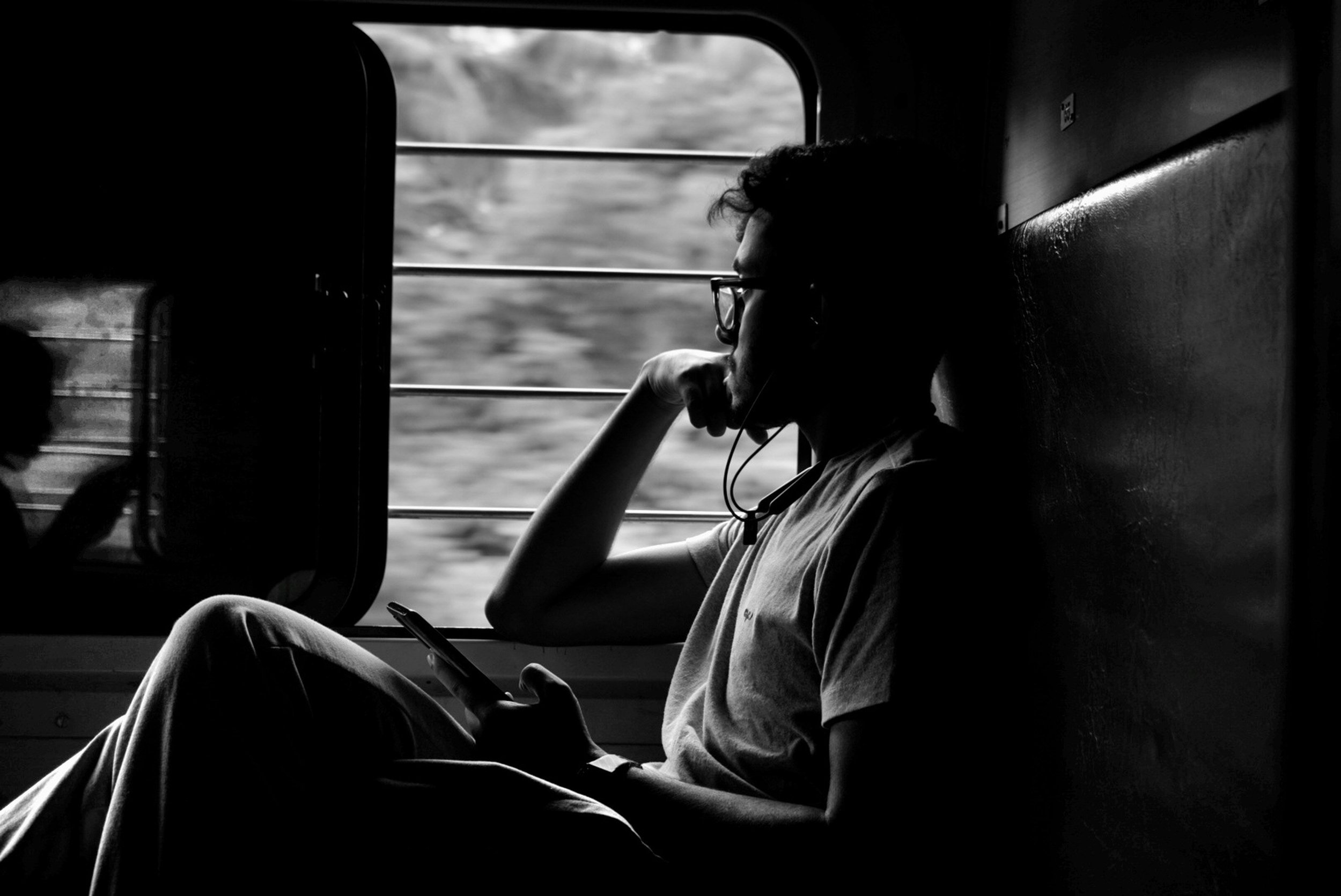 A black and white photo of a person with glasses sitting by a window on a train, looking at their phone with earphones in, relaxed and contemplative.