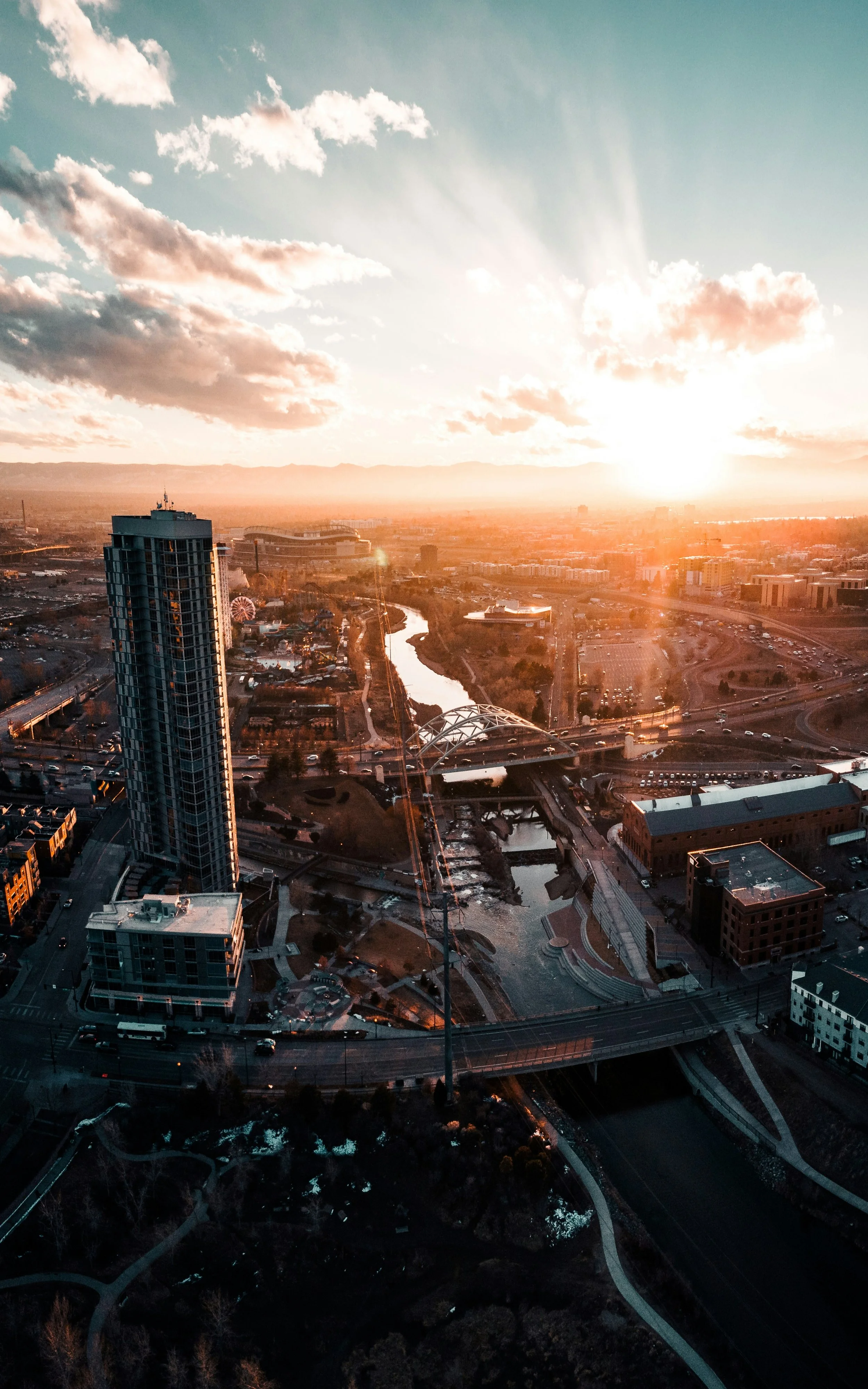 An aerial view of a city at sunset, featuring high-rise buildings, a river, and a park with winding pathways.