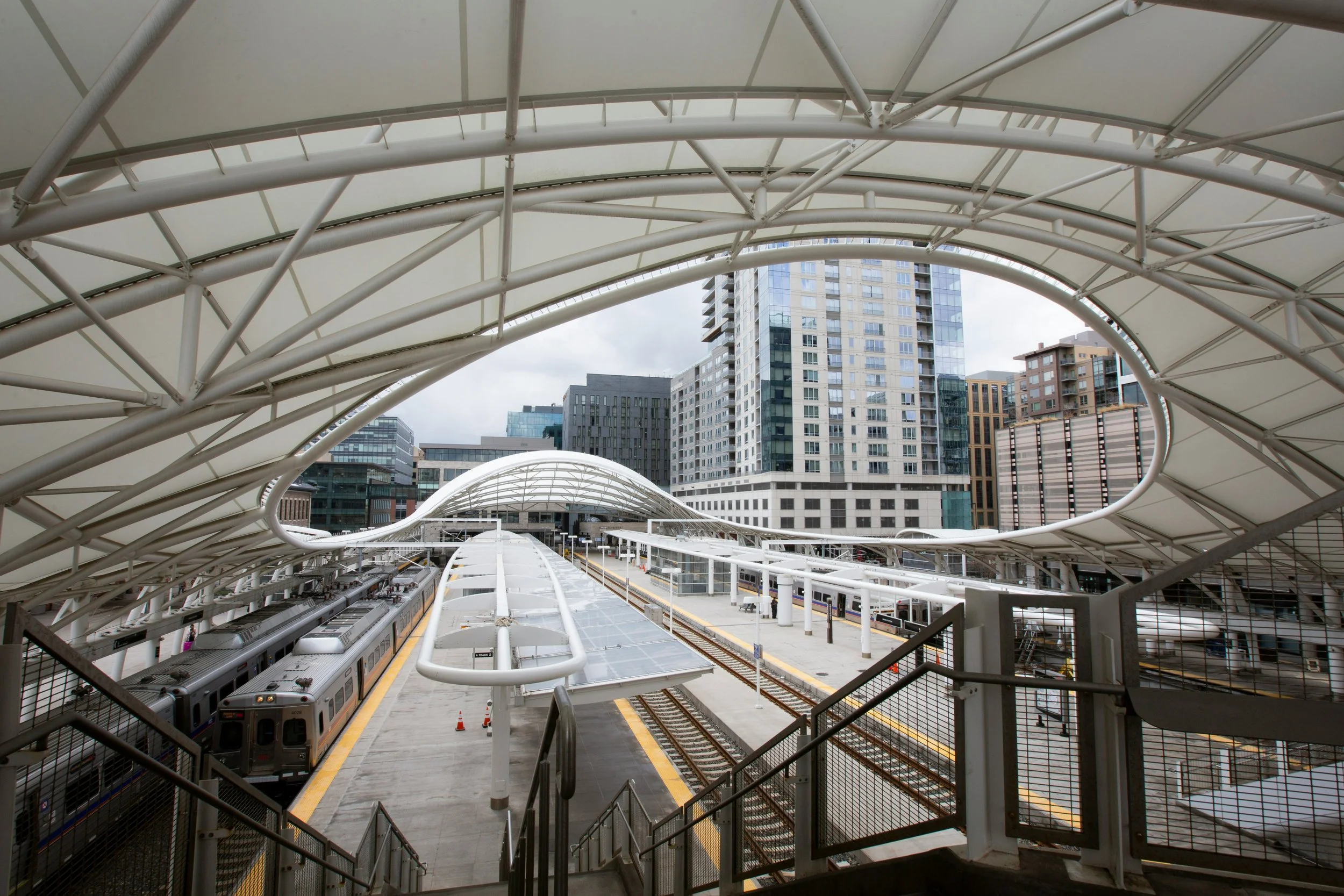 View of a modern train station with covered walkways, train tracks, and a train ready for departure, surrounded by tall city buildings.