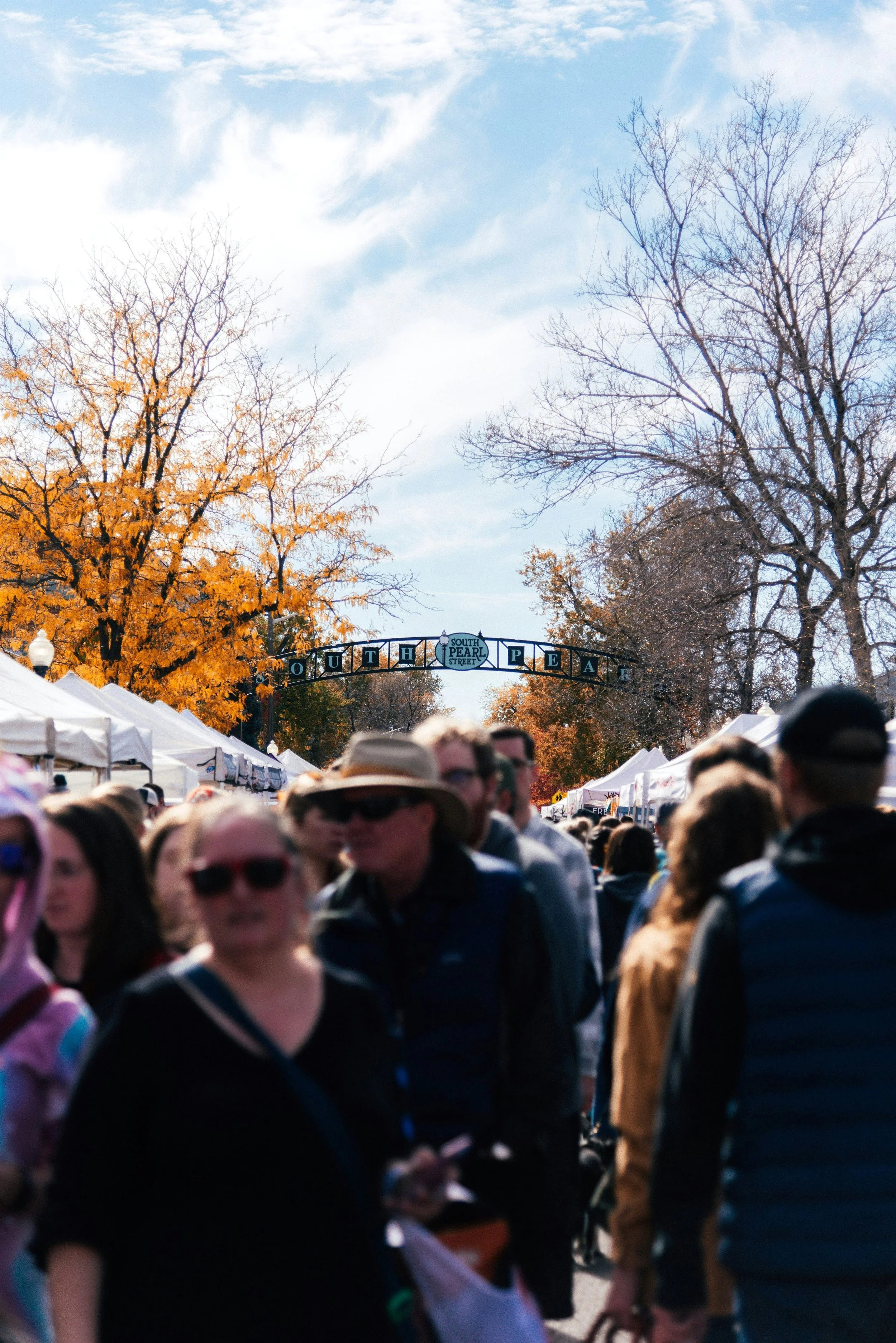 Crowd of people walking outdoors during autumn, with tents and trees with yellow and bare branches, and a street sign reading 'South Pearl Street' in the background.