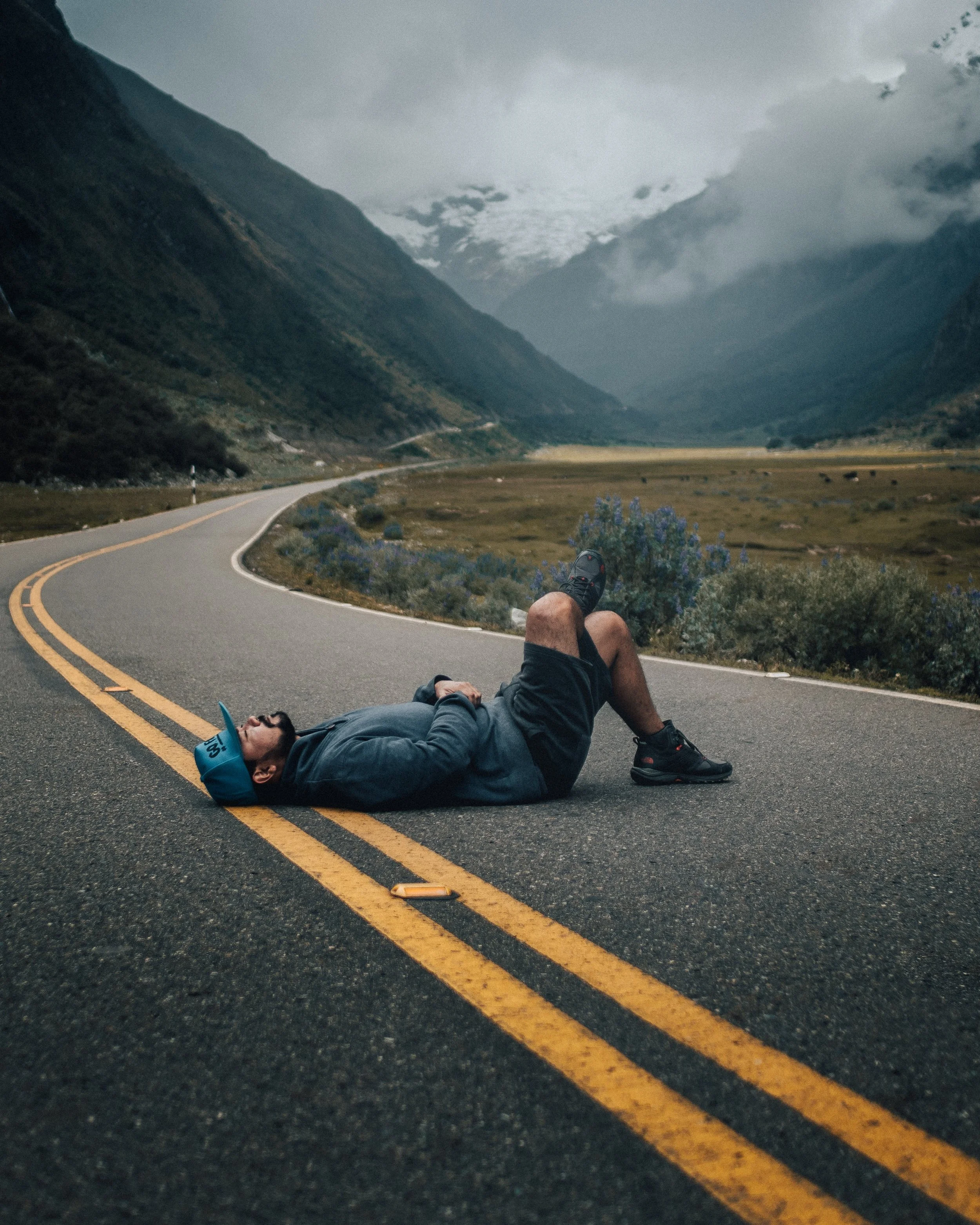 Person lying down on a winding mountain road with cloudy skies, mountains in the background, and sparse vegetation and flowers along the roadside.