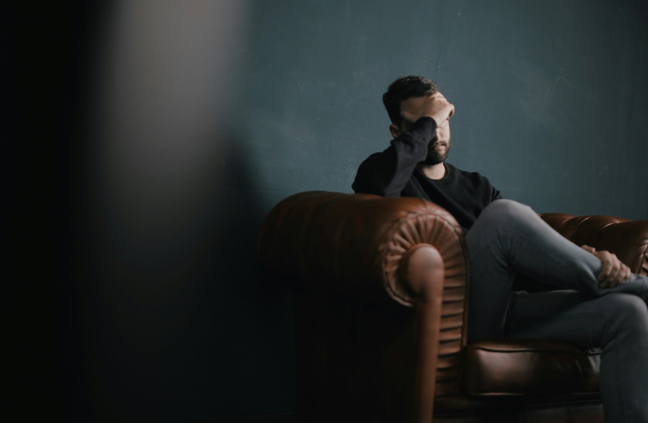 A man with dark hair and a beard sitting on a brown leather Chesterfield sofa, holding his forehead with one hand, against a dark teal wall.