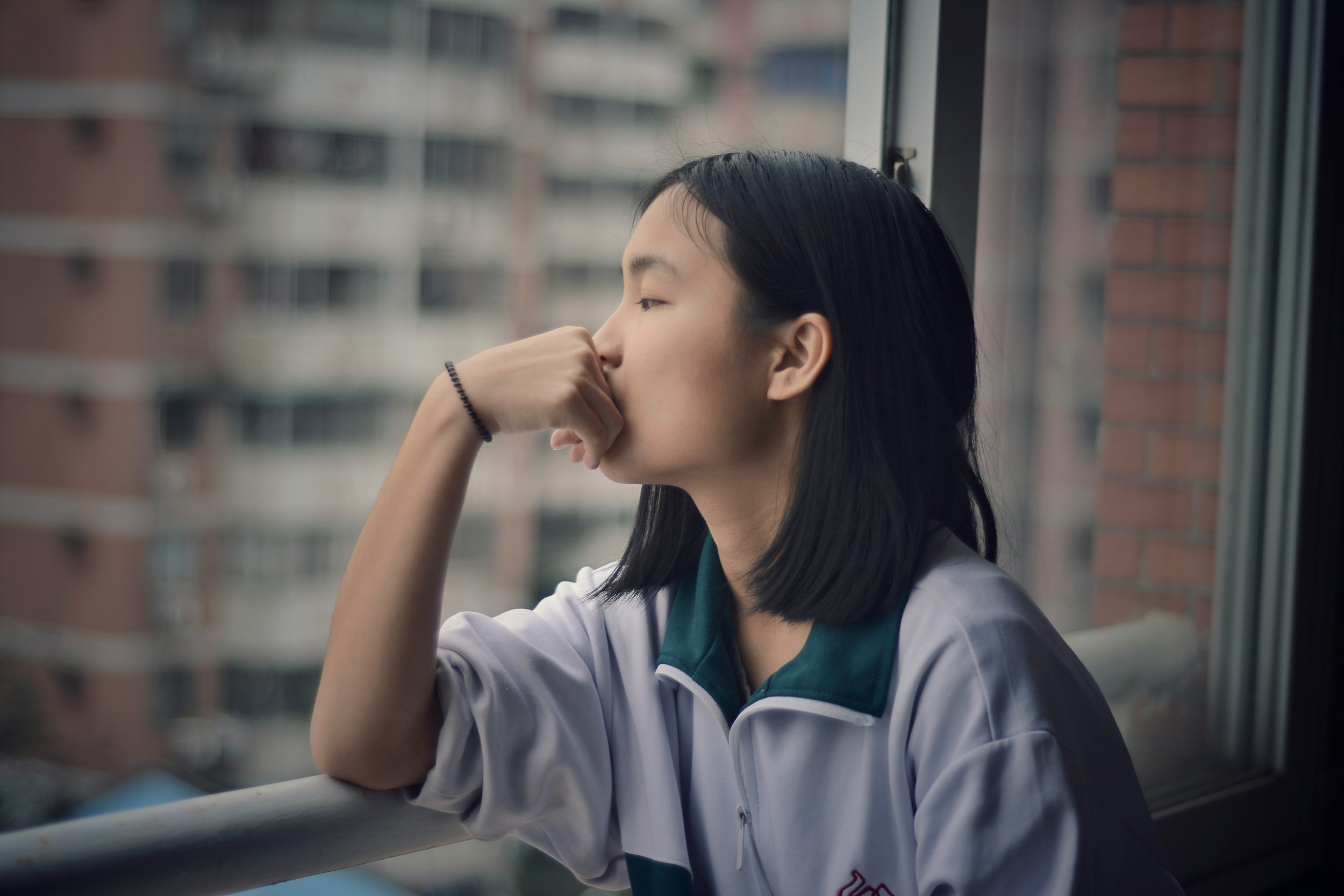 A young woman with dark hair and light skin, wearing a school uniform, sitting by a window with her hand covering her nose, looking contemplative outside at a cityscape with apartment buildings.
