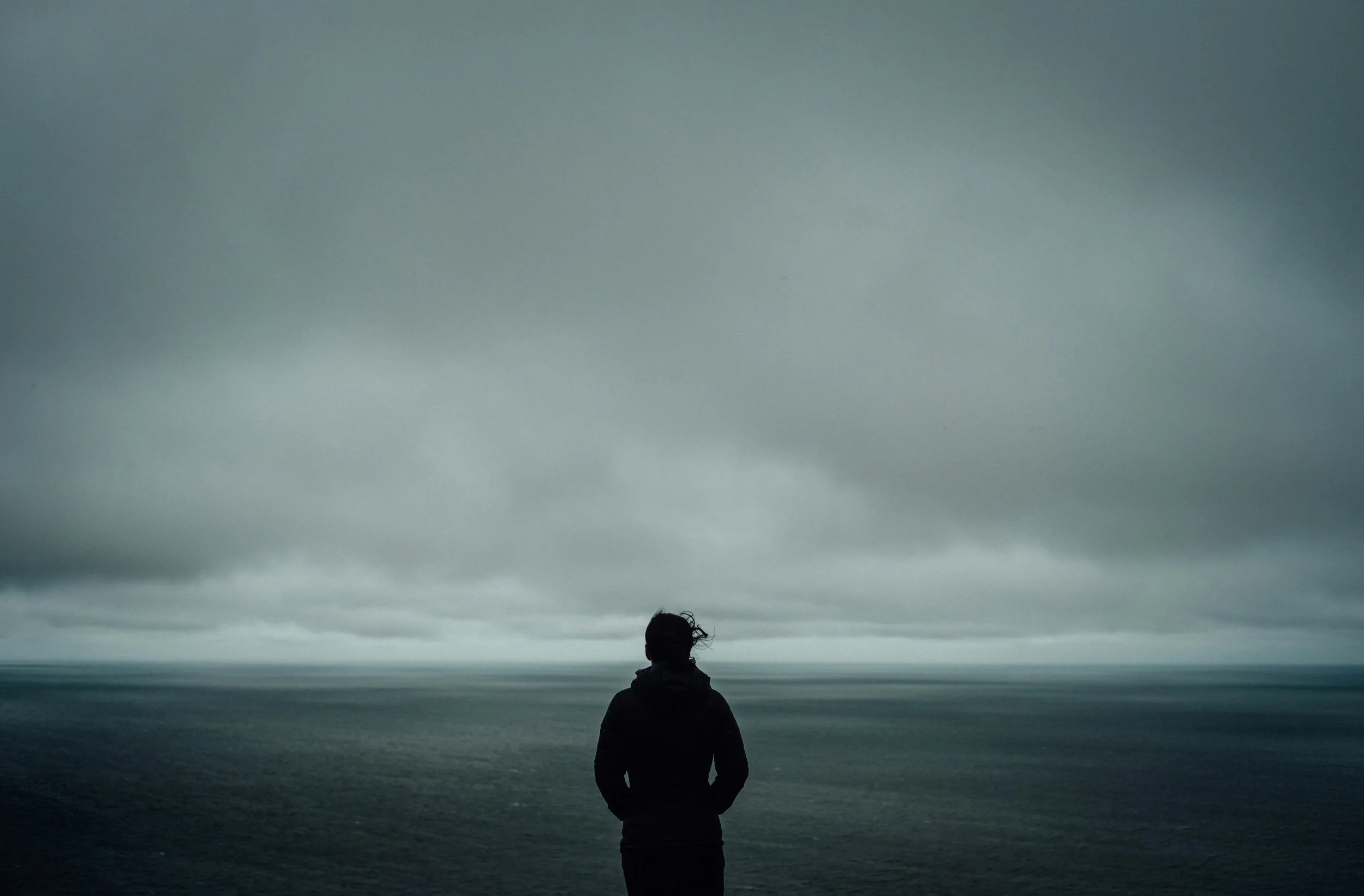A person stands on a beach facing the ocean, with cloudy sky overhead.