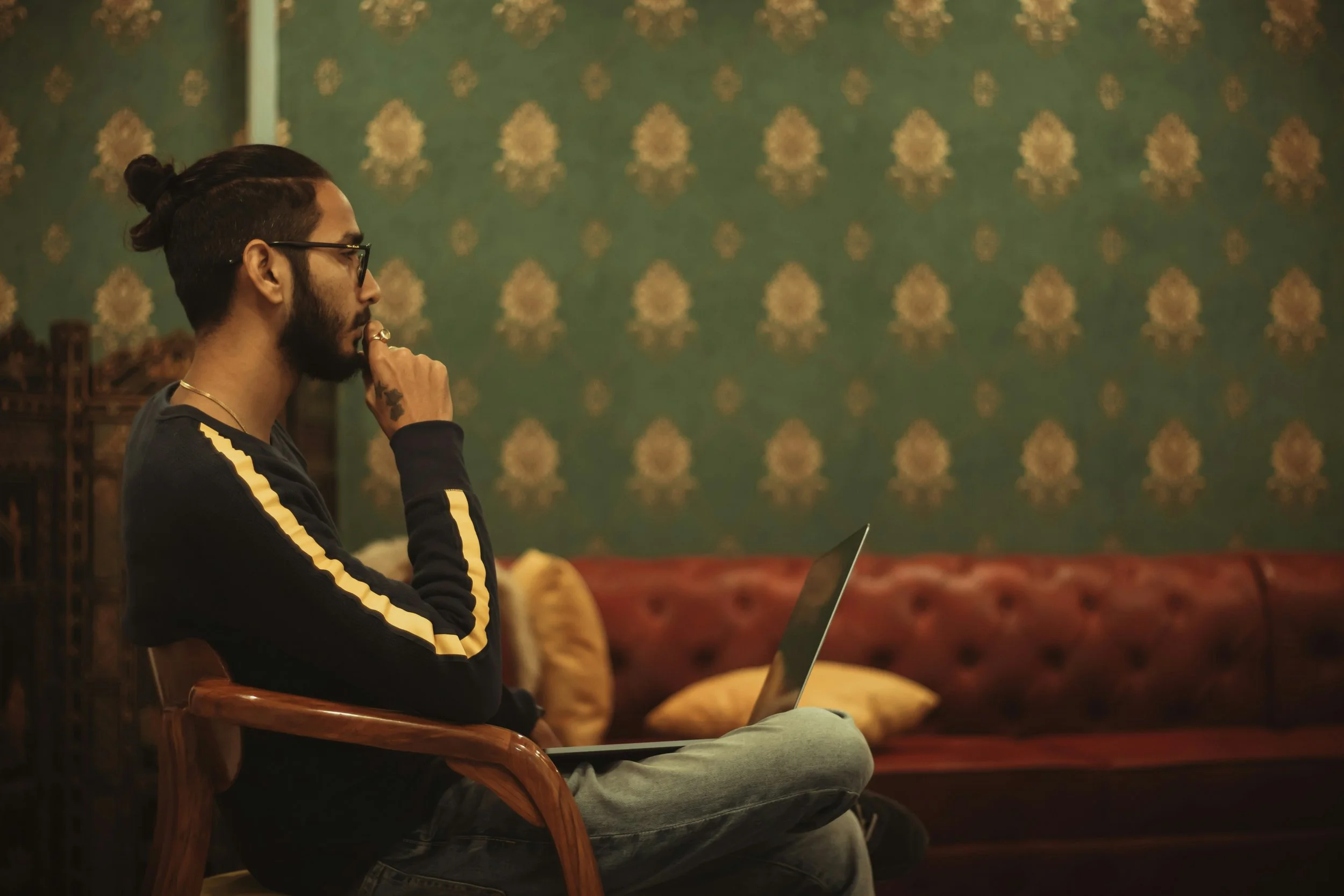 A man with glasses, a beard, and a man bun is sitting on a wooden chair, looking at a laptop, with his hand on his chin, in a room with vintage-style green wallpaper and cushions.