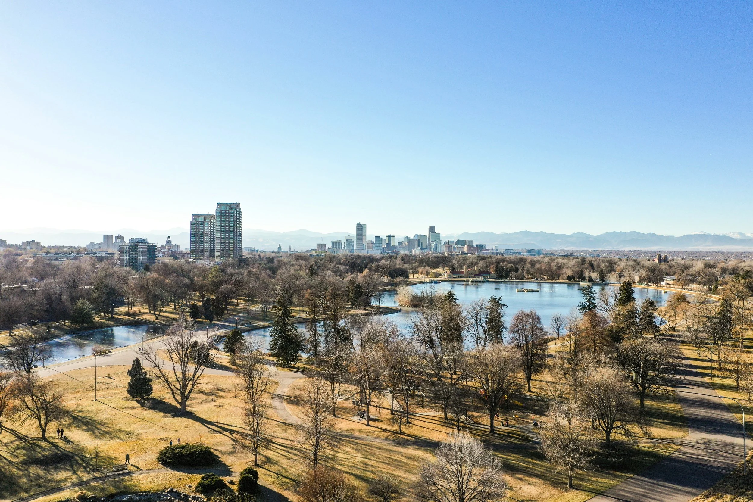 A city skyline with tall buildings in the distance, a body of water, and a park with trees and walking paths in the foreground on a clear day.