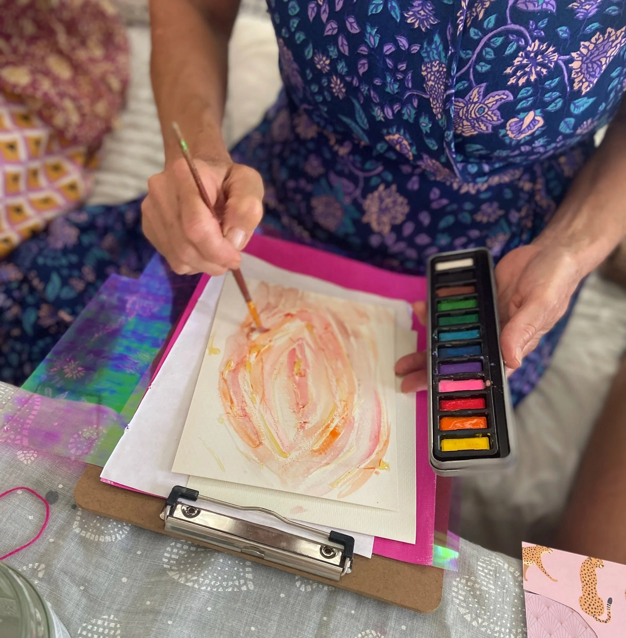 woman's hand painting a vulva shape with a set of watercolour paints in the other hand