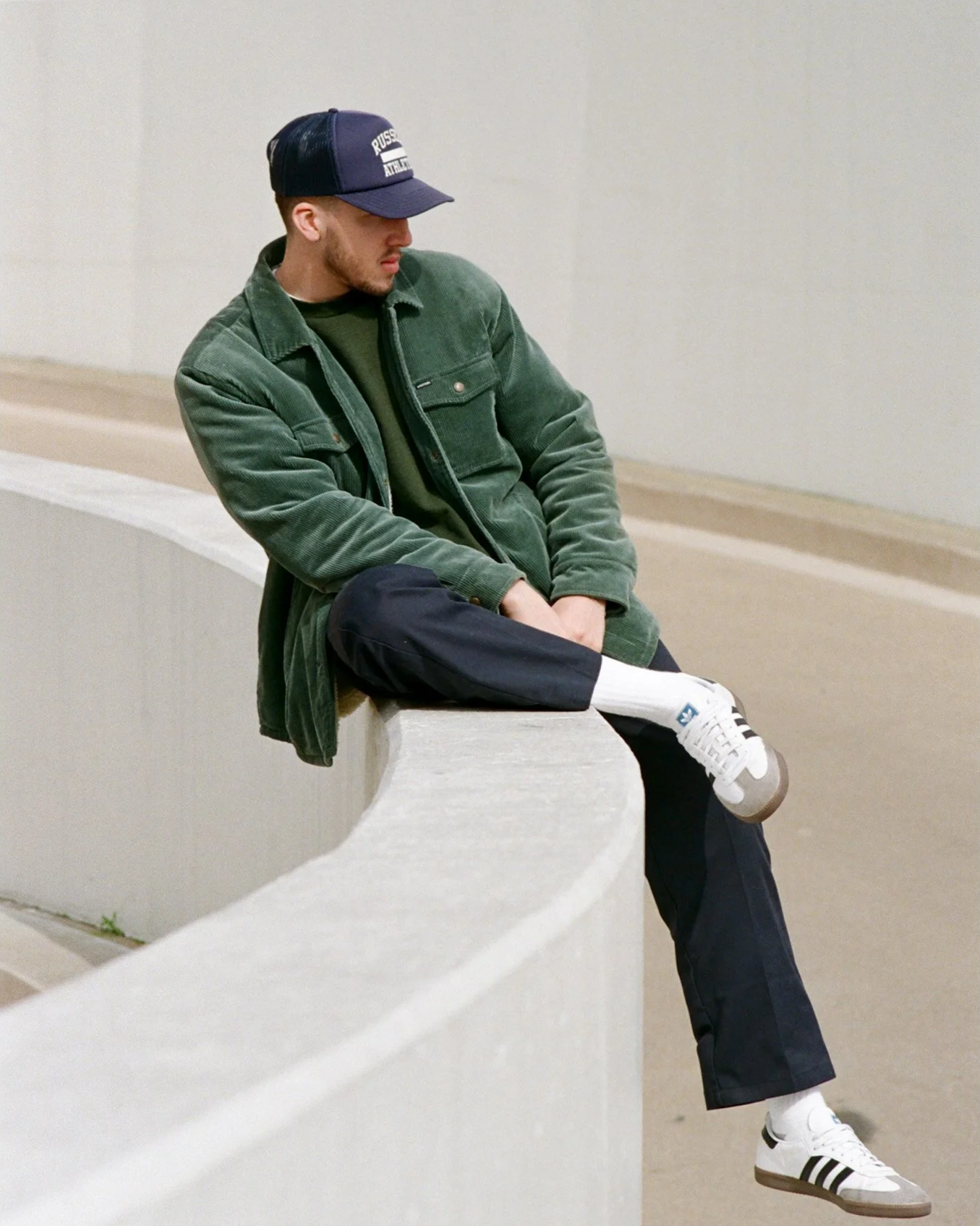 A man in a green jacket, black pants, and white sneakers with black stripes sitting on a white concrete ledge in an outdoor space with neutral-colored walls and a fence in the background.