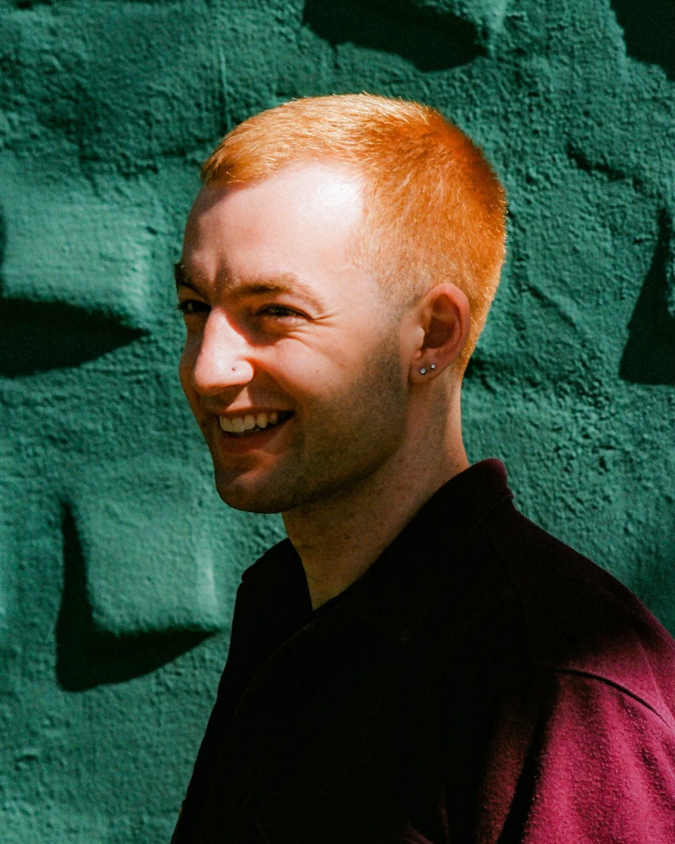 Smiling young man with short red hair and earrings standing in front of a textured green wall
