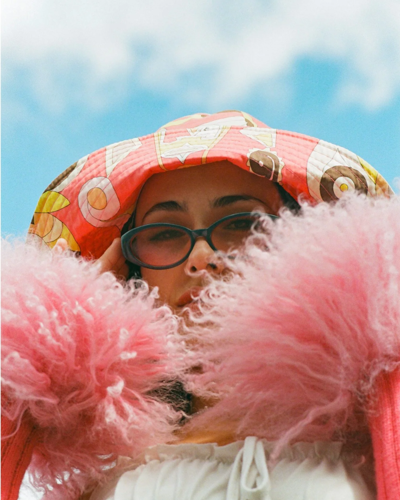 A woman wearing glasses and a colorful patterned hat, with pink fluffy sleeves covering her shoulders, looking down at the camera against a blue sky with some clouds.