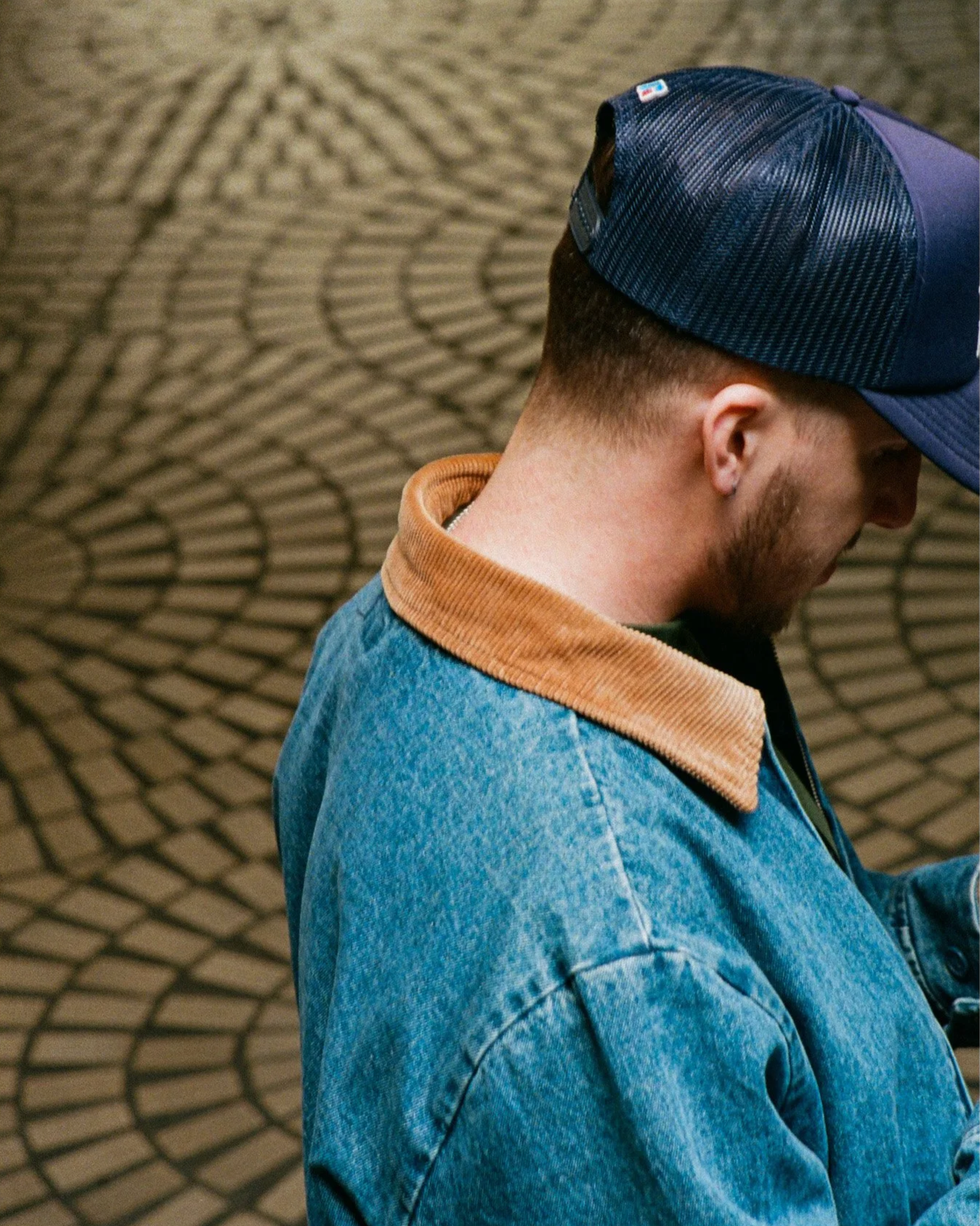 Side profile of a young man wearing a blue denim jacket, a tan collar, and a navy blue and black cap with a mesh back, standing on a cobblestone street.