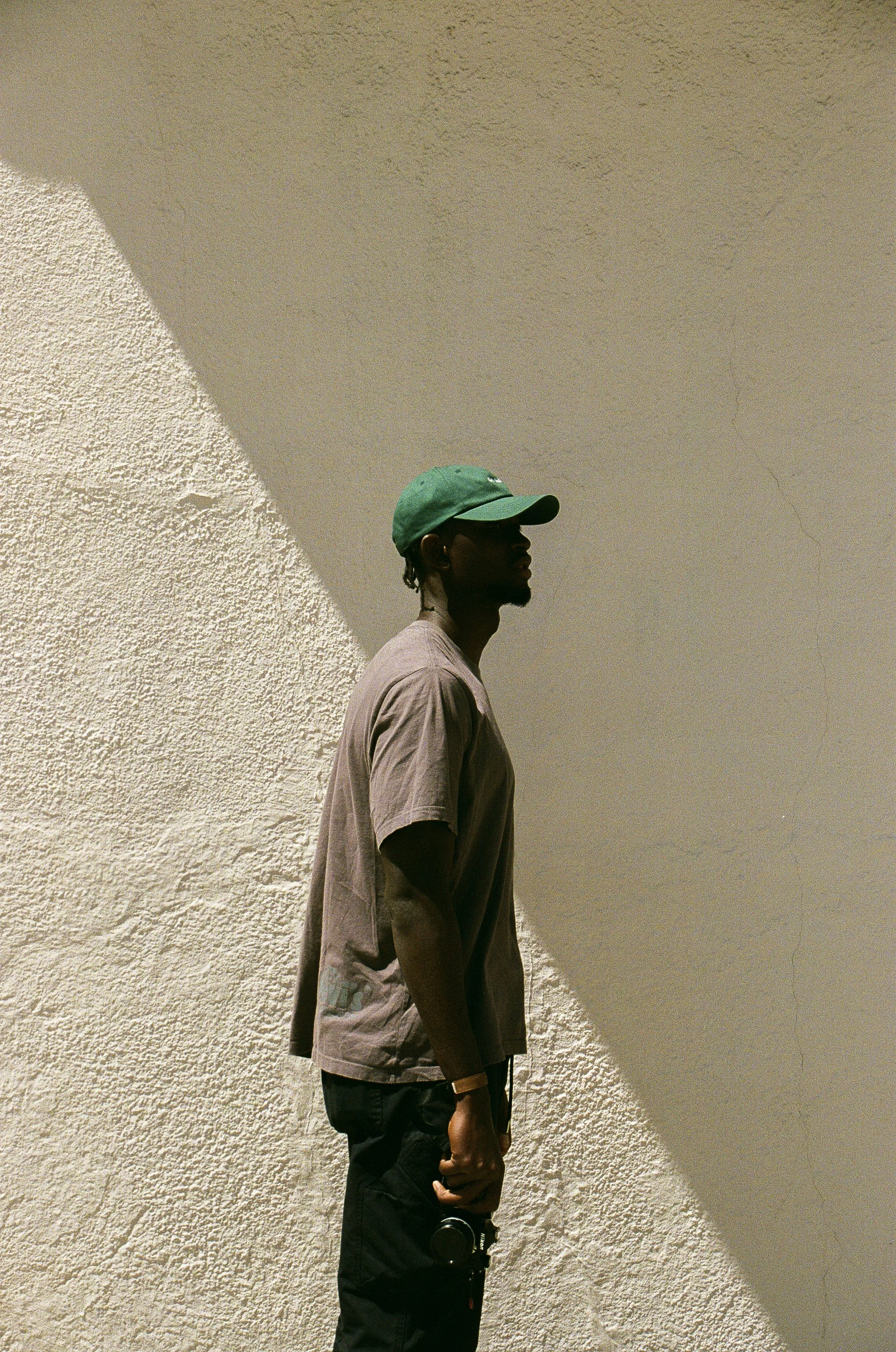 A young man standing against a textured wall with beige and white colors, wearing a green baseball cap, a loose brown T-shirt, and black pants, with a shadow cast on the wall behind him.