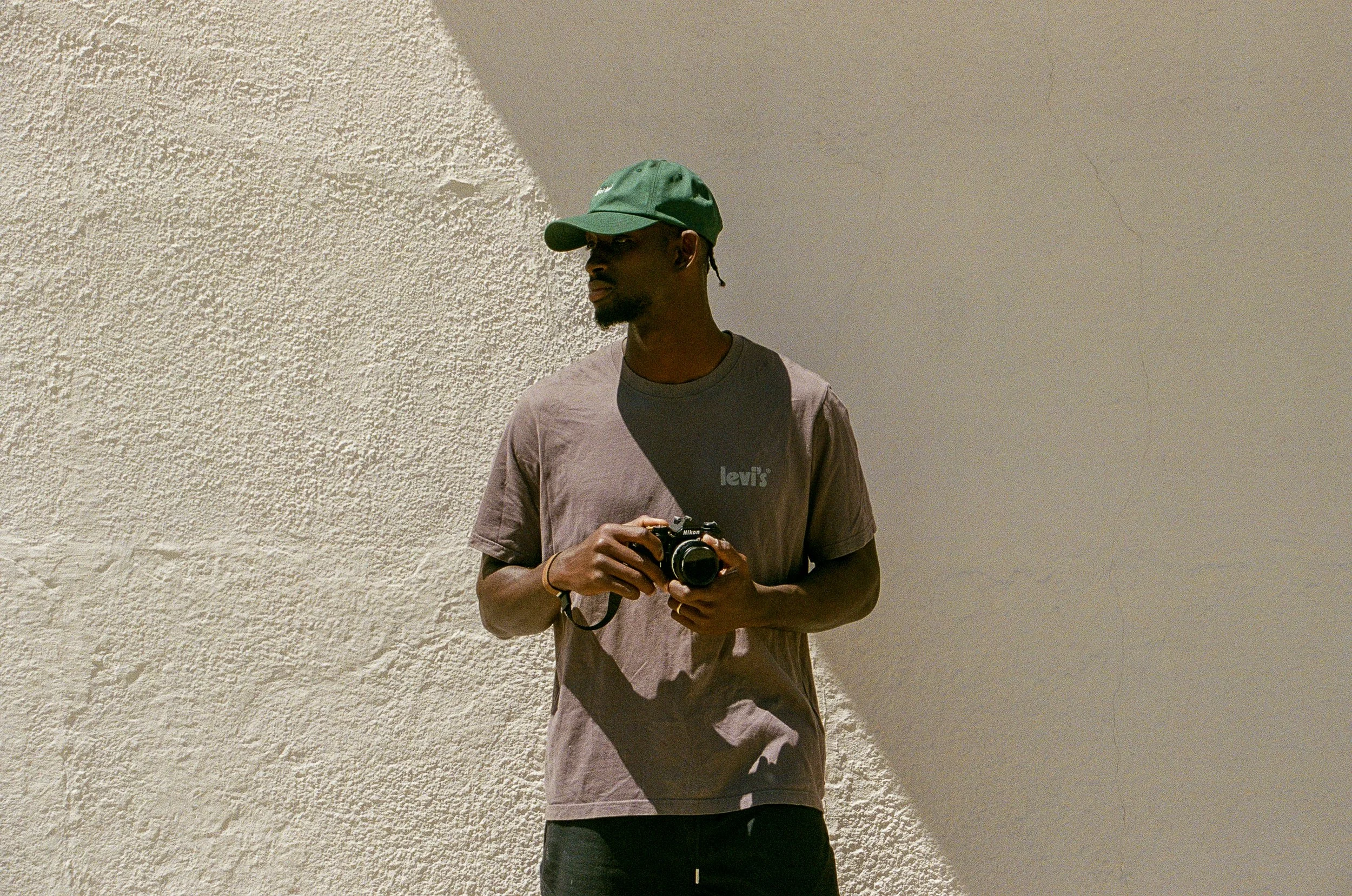 Young man wearing a green cap and a mauve Levi's t-shirt holding a camera standing against a textured off-white wall with partial shade.