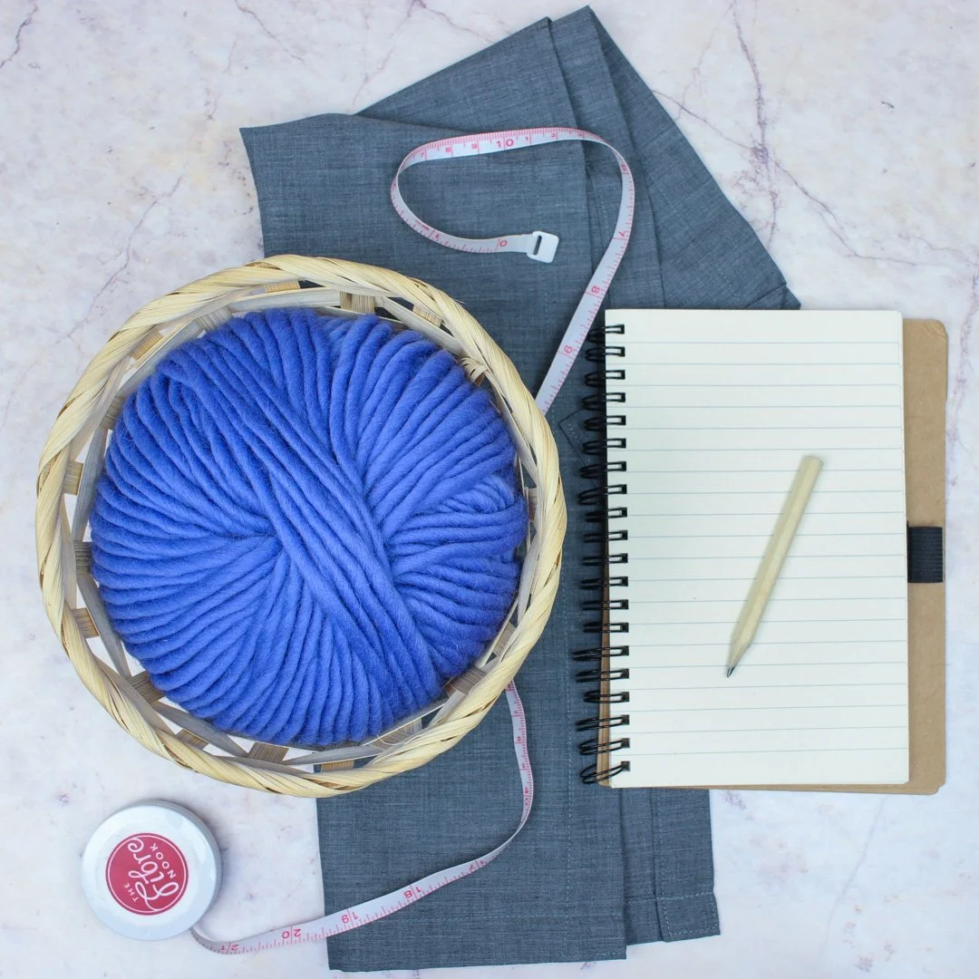 Flatlay showing a ball of &Make Super Bulky in shade potion, a notebook and pencil and a tape measure on a marble effect background.