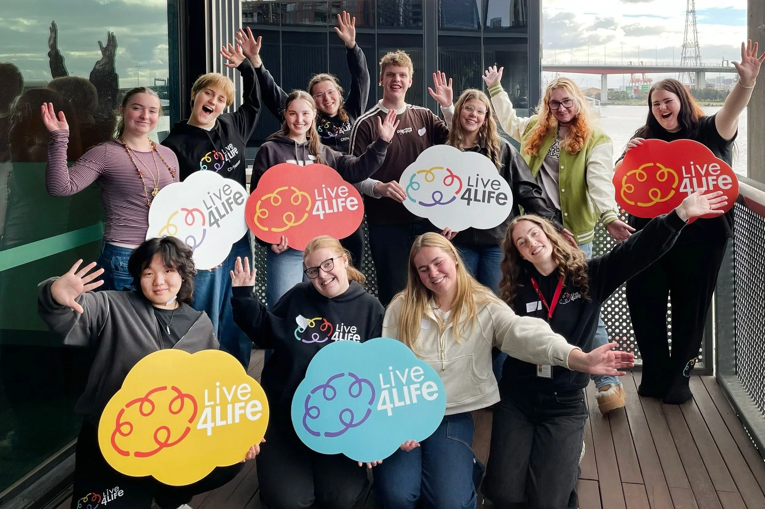 A group of young people laughing and holding Live4Life signs with Melbourne Docklands in the background