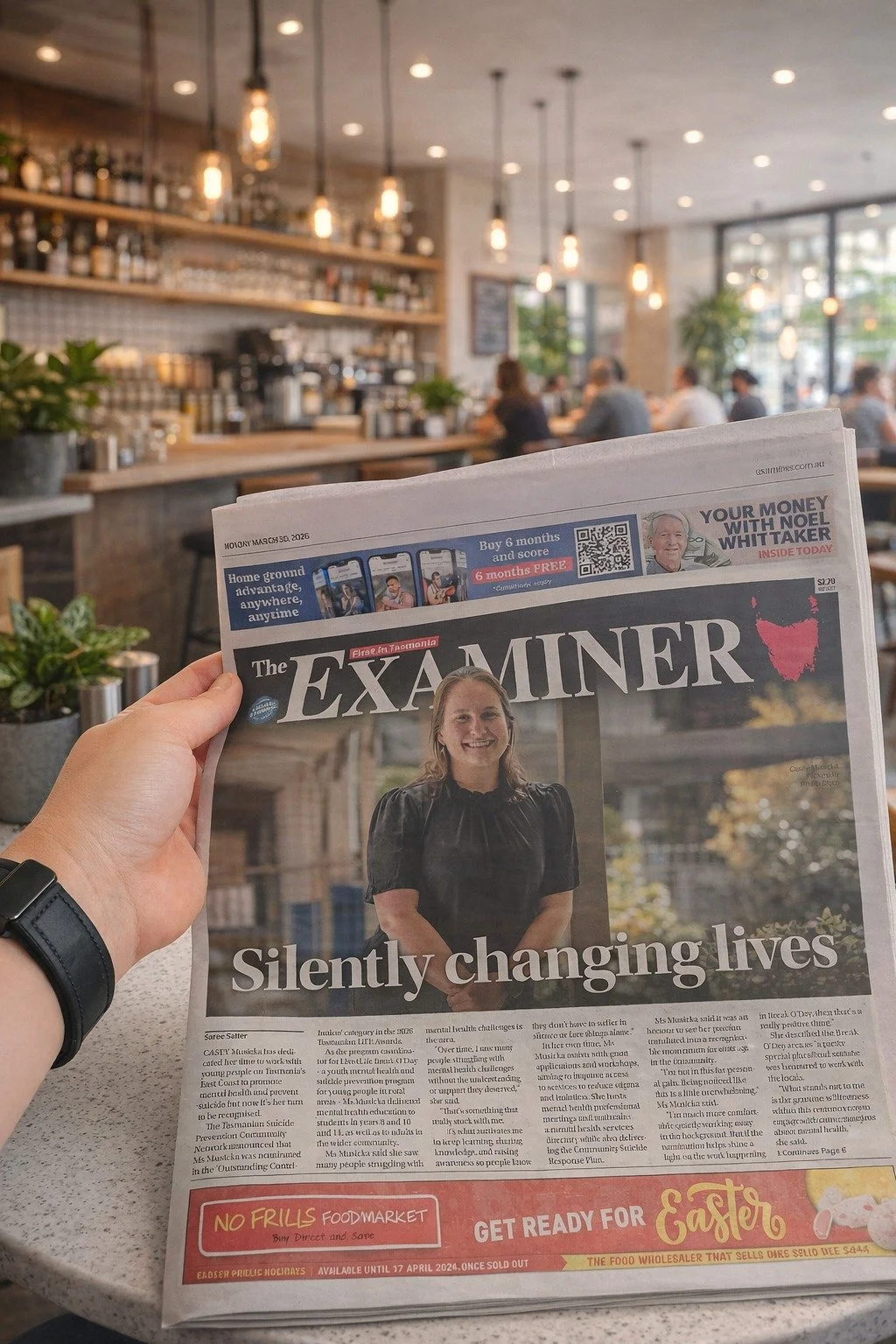 A hand holding up the front page of the Launceston Examiner with a cafe interior in the background