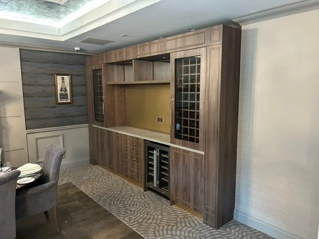 A built-in wooden entertainment unit with glass-paneled cabinets, a countertop, and a small wine cooler in a dining room with textured wall and carpeted floor.